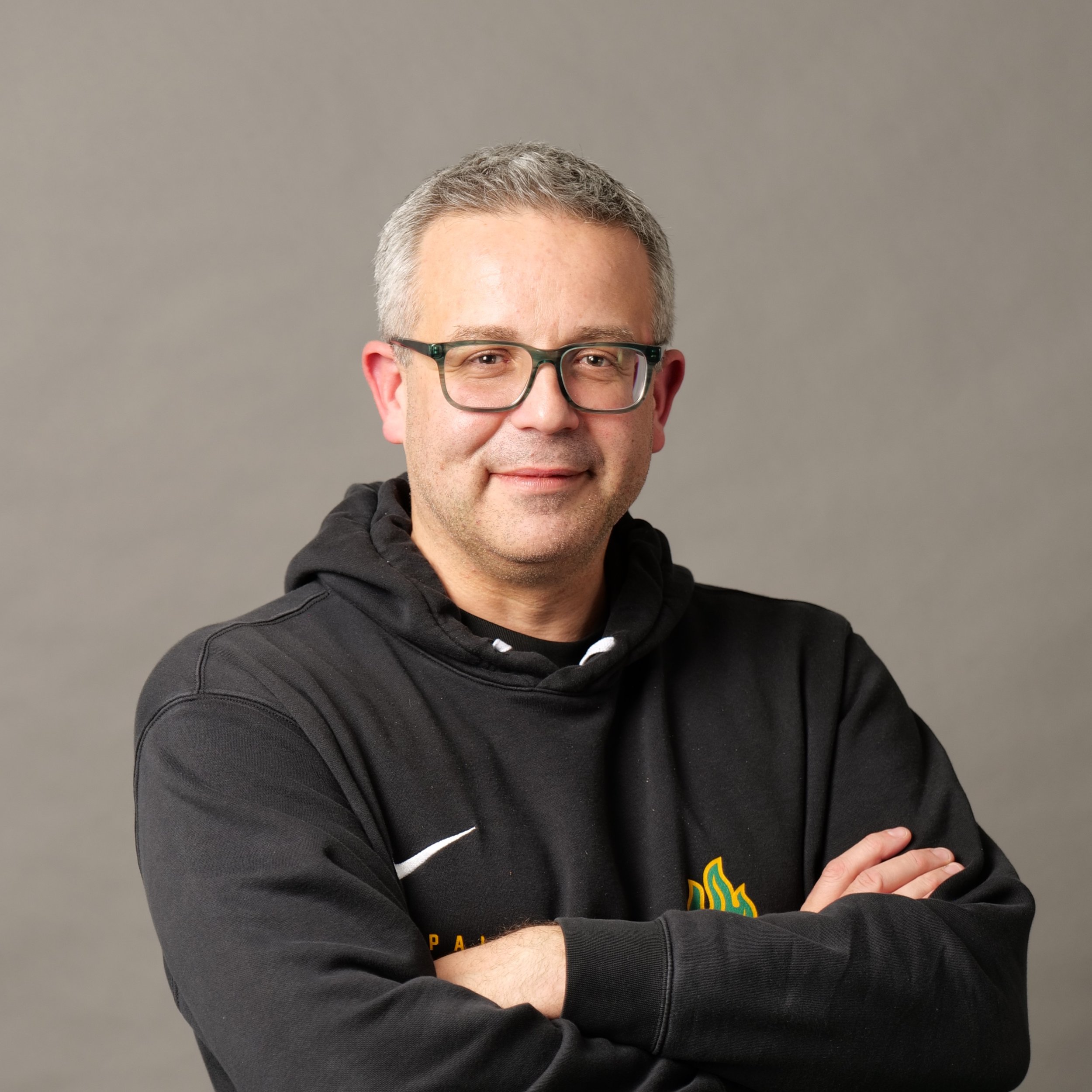 Andrea Zanolin, coach di basket, capelli corti e grigi, sorridente, che indossa una felpa di pallacanestro ponzano, in posa con le braccia incrociate, sfondo grigio.