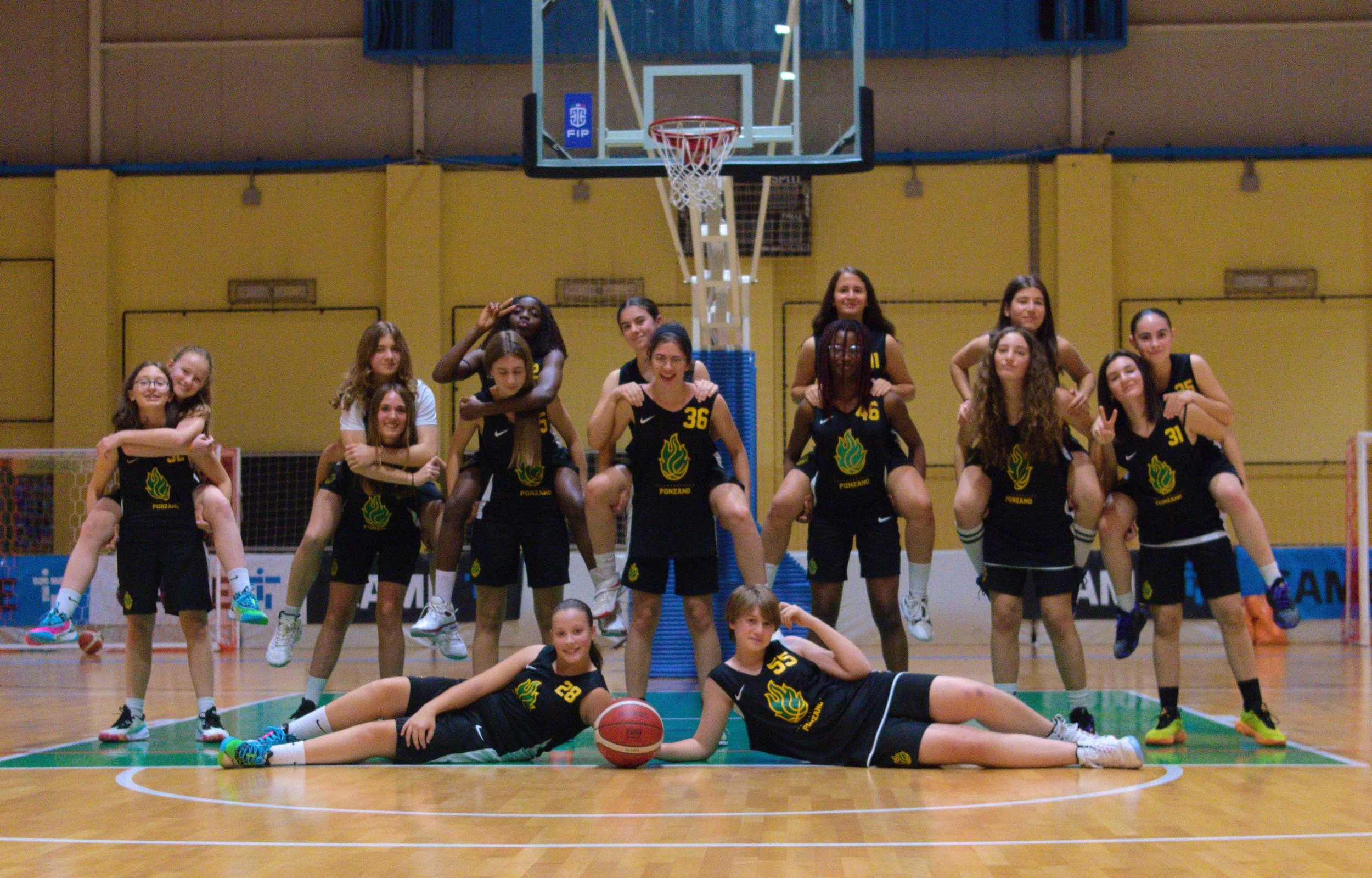 Squadra di under 14 femminile di basket in posa sul campo in palestra, alcune sono in piedi e altre sono sdraiate in prima fila con una palla da pallacanestro.