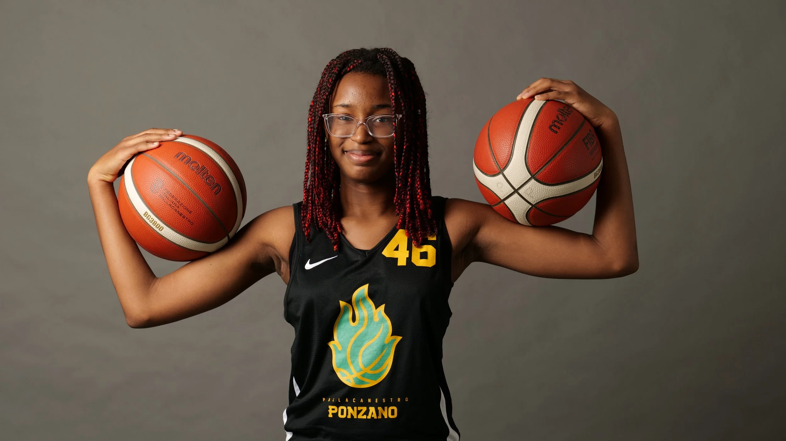 Una giocatrice di basket dell'u14 femminile con i capelli con riccioli rossi e neri, indossa una maglietta nera con il numero 45 e il logo di pallacanestro Ponzano, e tiene due palloni da basket, uno su ogni spalla, davanti a uno sfondo grigio.
