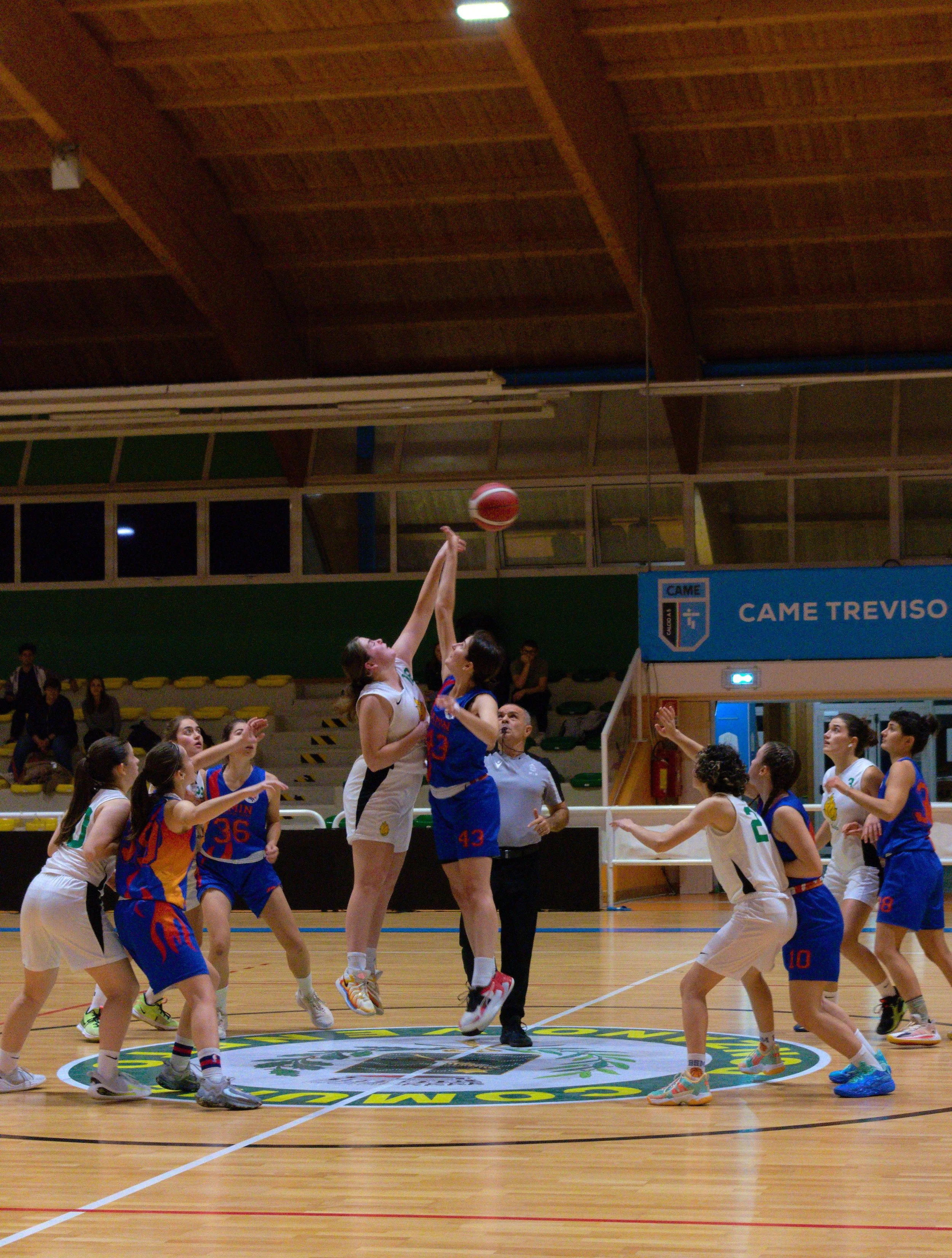 Partita di basket femminile tra due squadre di giovani, con due giocatrici che si contendono il rimbalzo durante il salto.