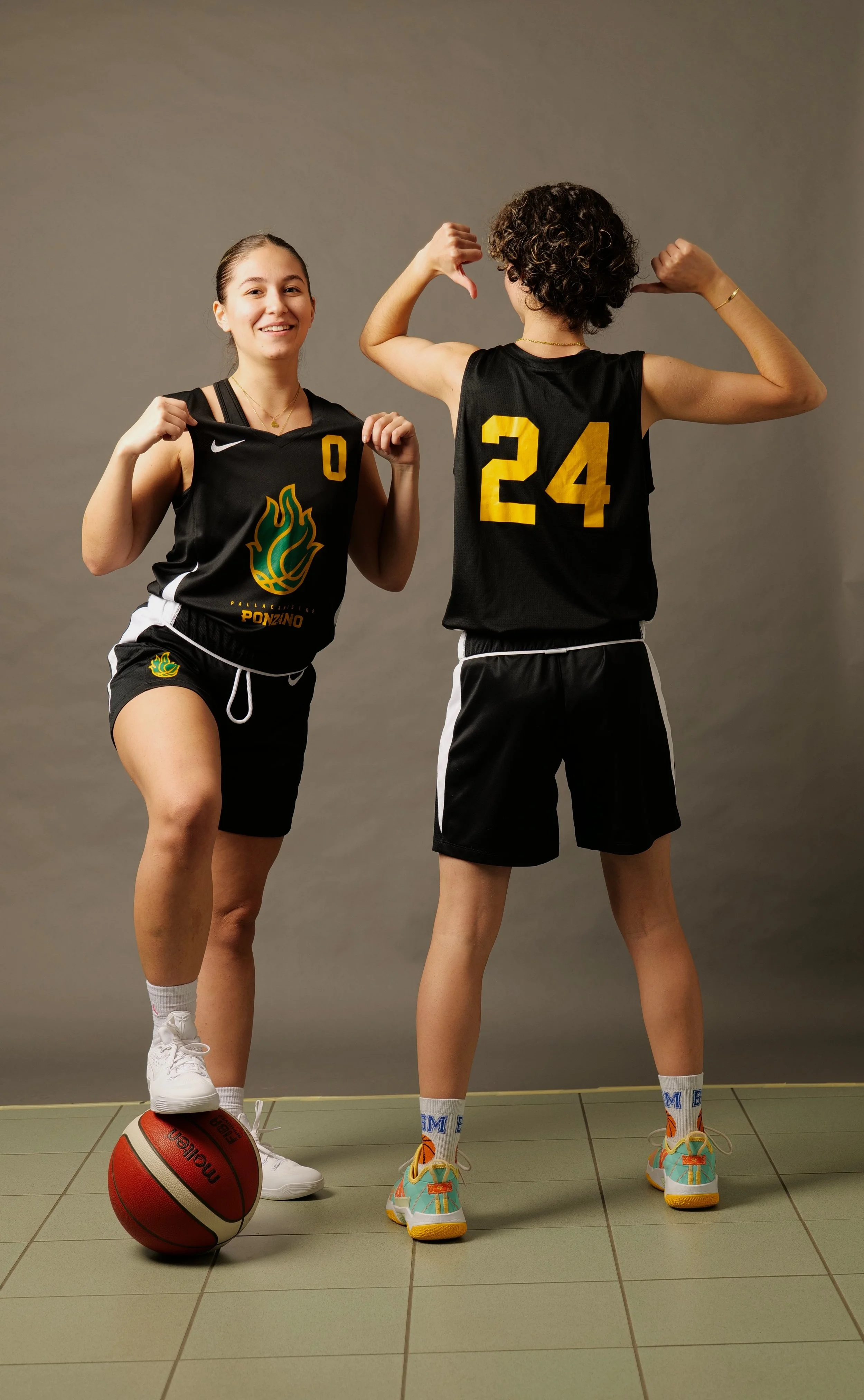 Due ragazze della promozione femminile in divisa da basket della pallacanestro ponzano posano, una con una palla da basket e l'altra che mostra i muscoli, contro uno sfondo grigio.