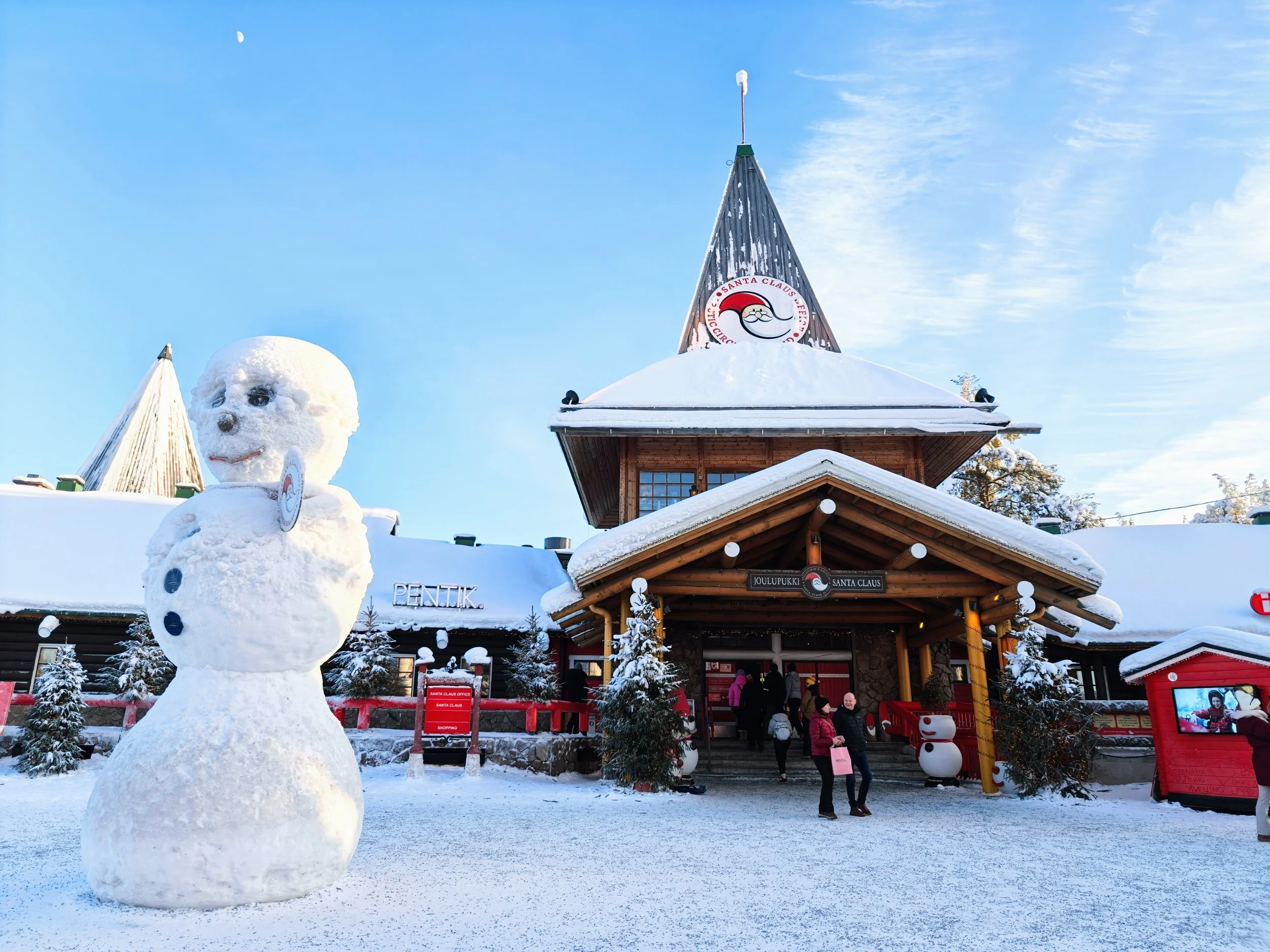Snowman with sunglasses and buttons standing outside Santa Claus village in Lapland, Finland, in winter, with wooden buildings and a snow-covered landscape.
