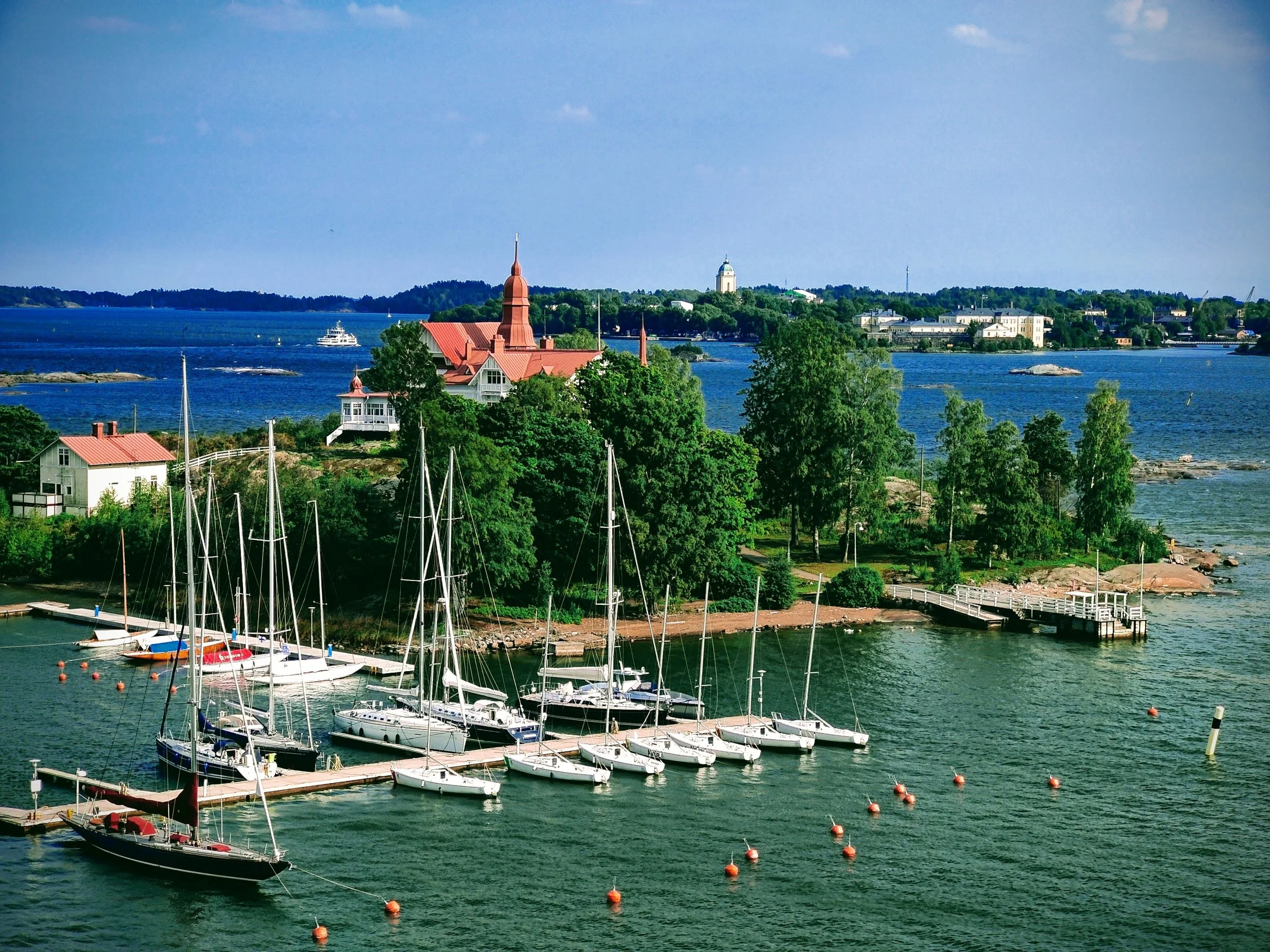 A scenic waterfront view of sailboats docked at a marina, with lush green trees and historic buildings on a small island, and a large body of water extending to the horizon under a partly cloudy sky.