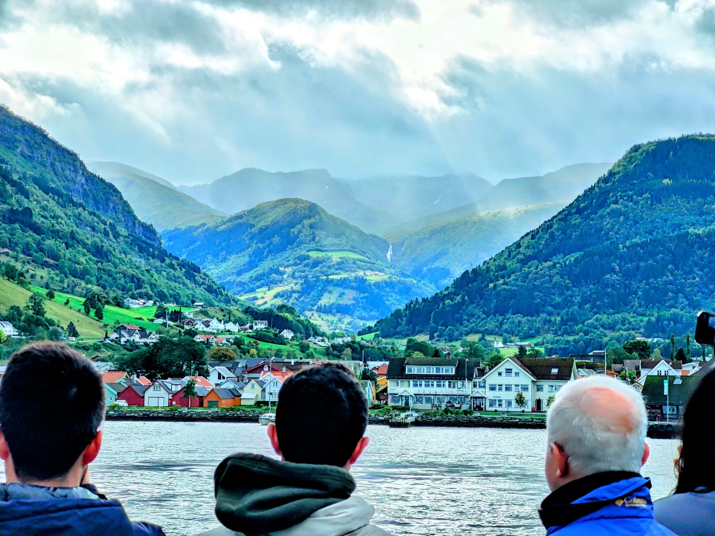 Four people sit by a body of water watching a scenic view of a small town on the lake, with colorful houses and lush green hills and mountains in the background under a partly cloudy sky.