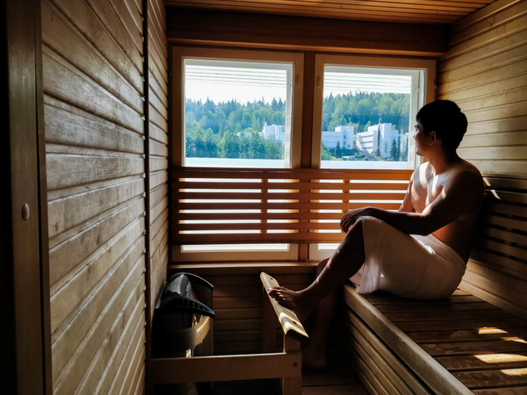 A person sitting inside a wooden sauna, looking out the window at a scenic view of trees and buildings.