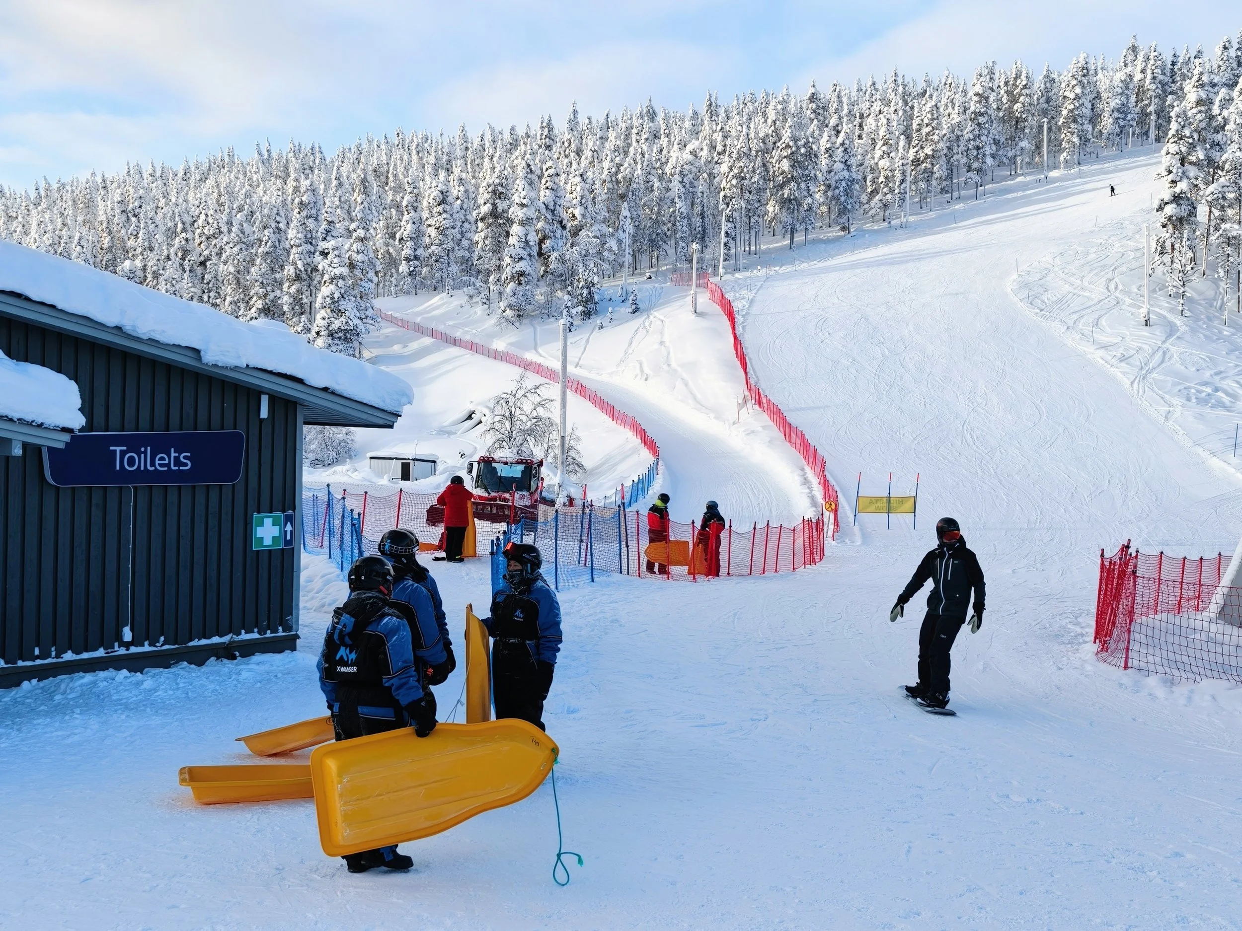 Ski resort with snow-covered slopes, trees, and people in winter gear preparing for skiing and snowboarding near a building with a 'Toilets' sign.