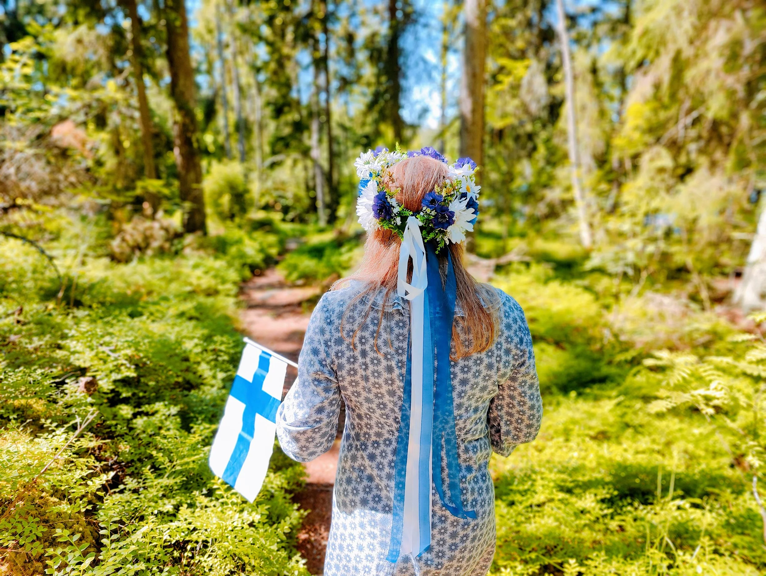 A woman with long, light brown hair wearing a flower crown and traditional blue and white dress, walking in a forest during daytime, carrying a Finnish flag.