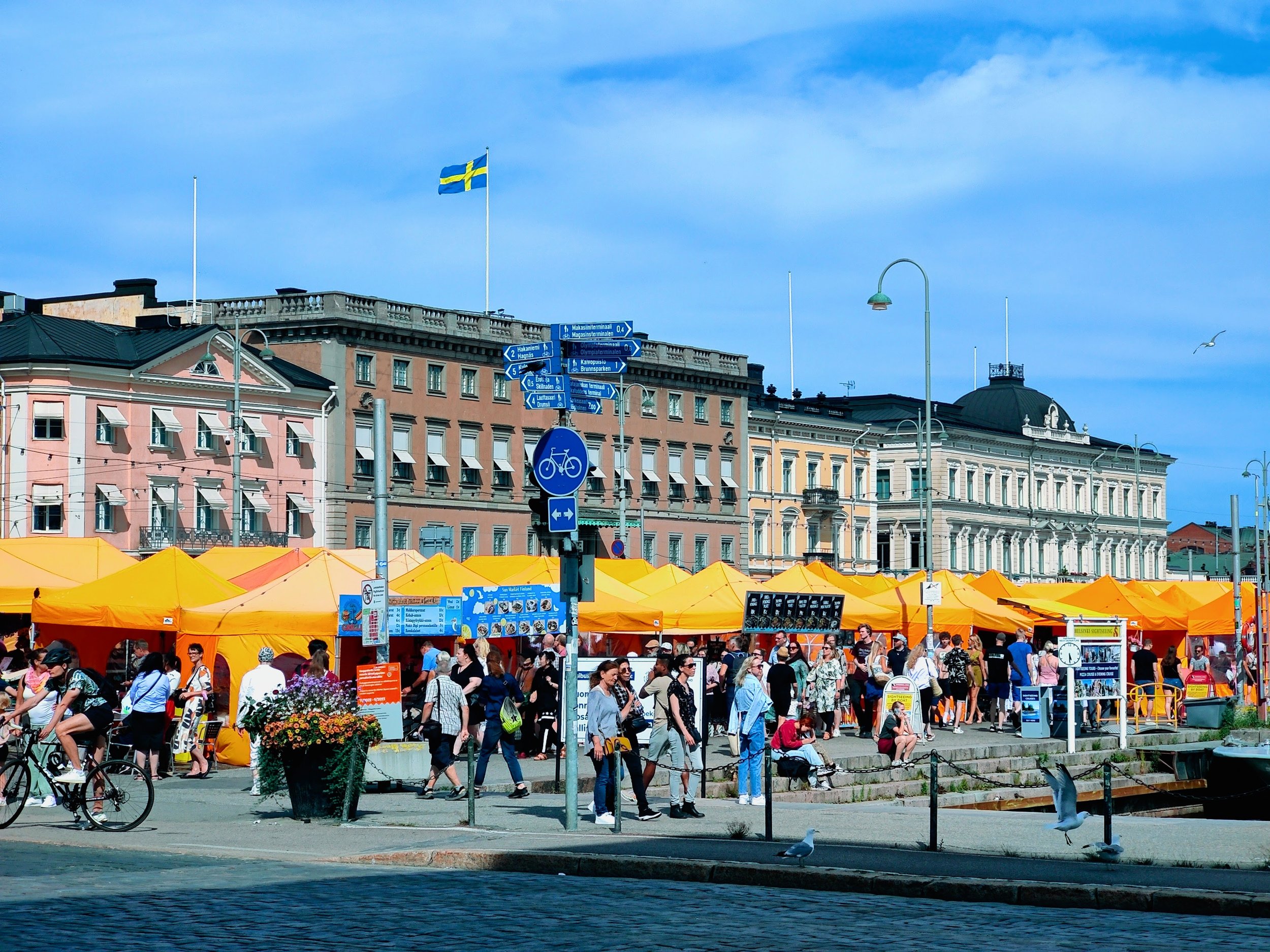 An outdoor market with yellow tents, people walking, and a bicycle, set against colorful historic buildings under a blue sky with Swedish flag.