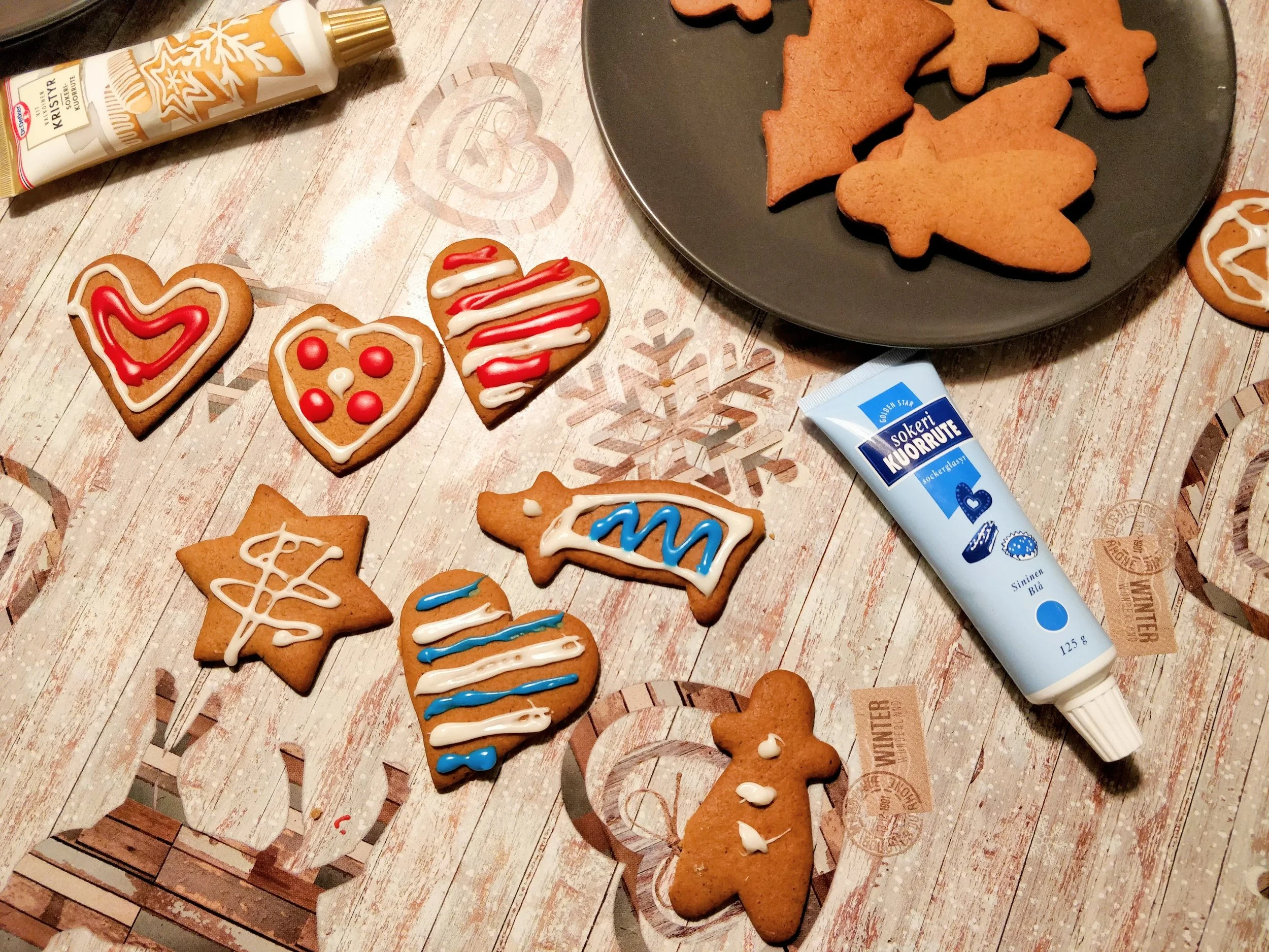 Assorted decorated gingerbread cookies on table, some with white, blue, red icing in various patterns, and a small gingerbread man cookie with white dots. Plate of plain gingerbread cookies in the background, and a tube of white icing on the table.