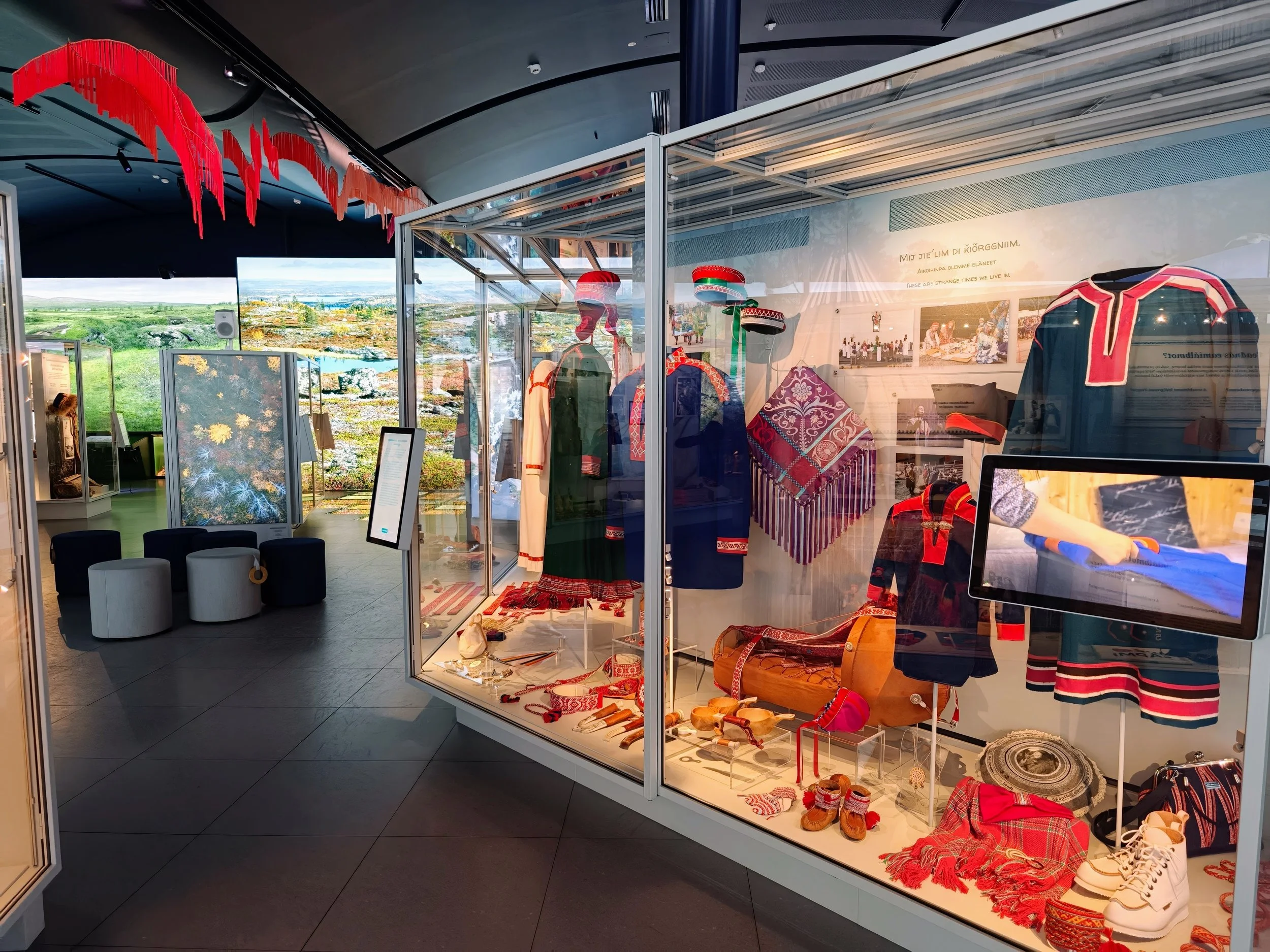 Museum display case with traditional artifacts including clothing, textiles, shoes, and accessories, alongside informational panels and a large screen showing related footage. ADA seating and digital exhibits visible in the background.