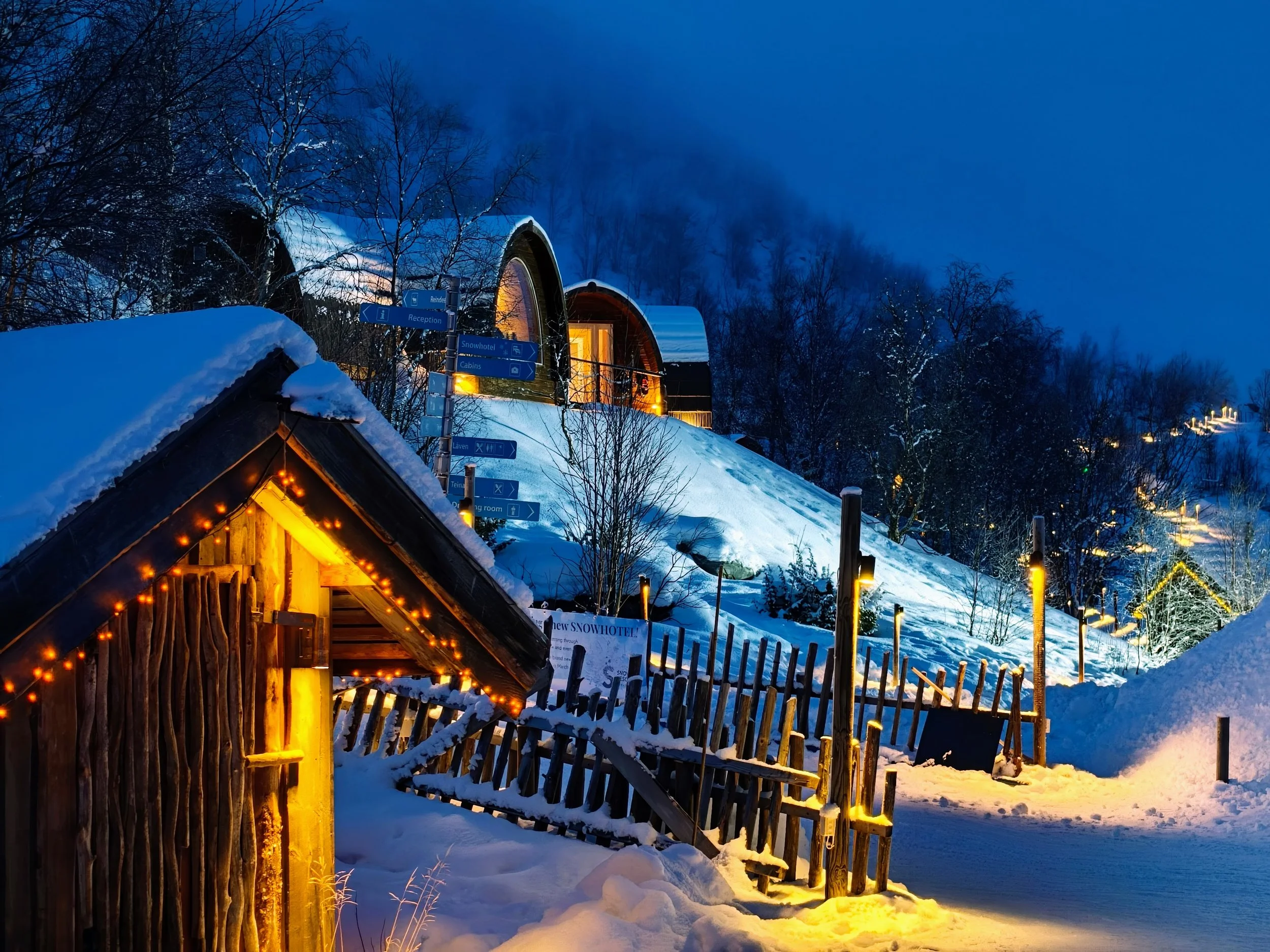 Snowy hillside with illuminated wooden cabins, a small hut with string lights in the foreground, and a modern chalet-style building on the slope at dusk, with snow-covered trees and a cloudy sky in the background.