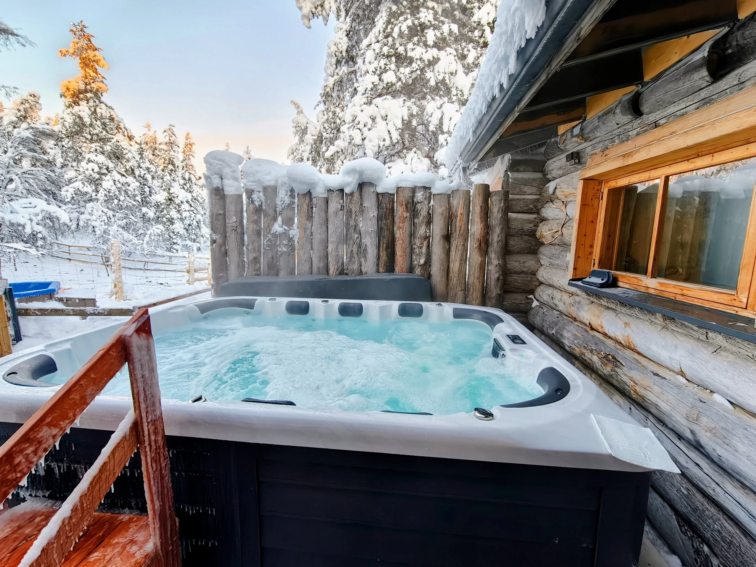 Hot tub on a wooden deck attached to a rustic log cabin, surrounded by snow-covered trees and a wooden fence, with snow on the tub's edge and a small window on the cabin wall.
