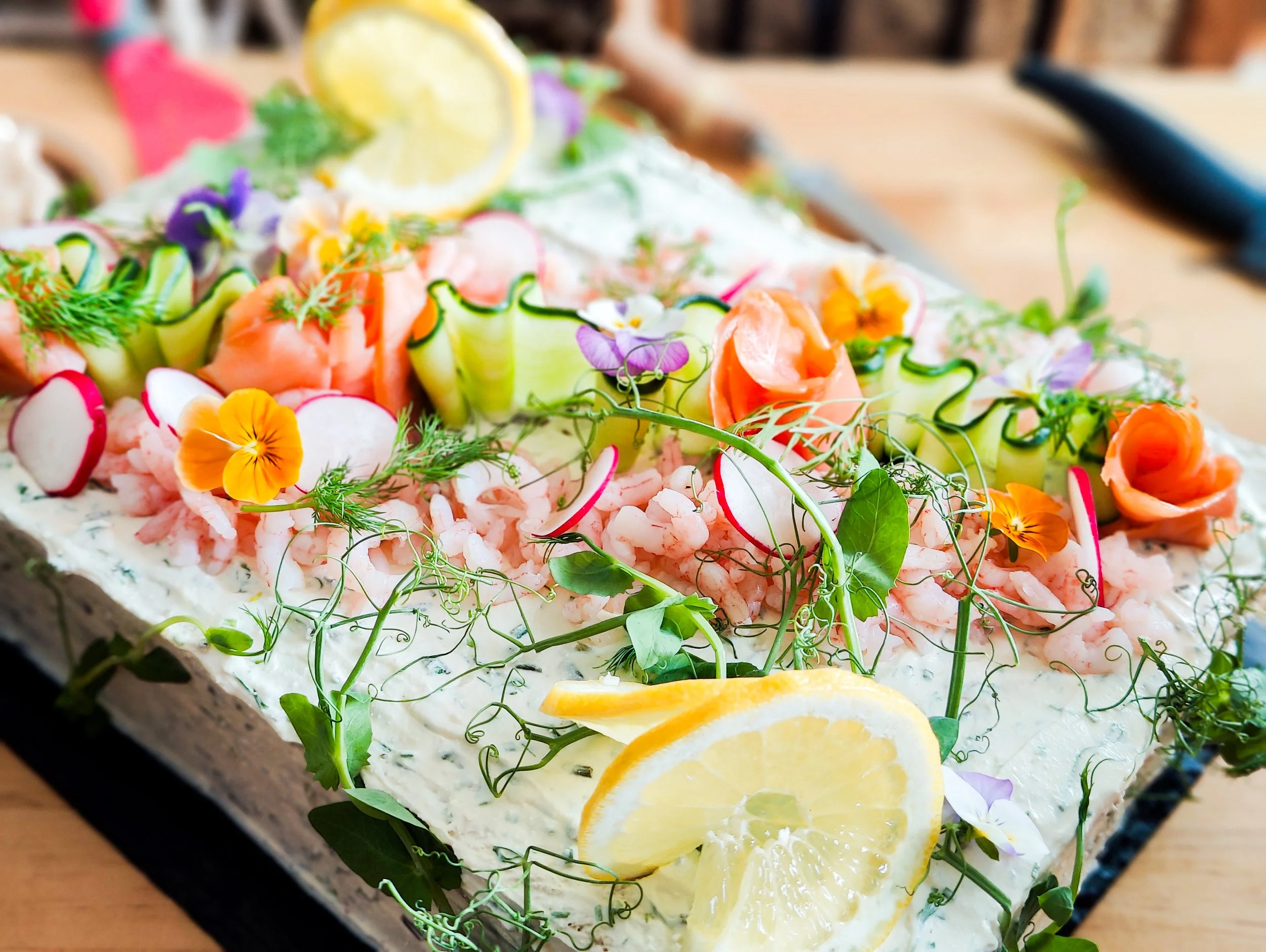 A colorful seafood platter with lemon slices, radish slices, cucumber ribbons, edible flowers, and fresh herbs on a bed of creamy sauce.