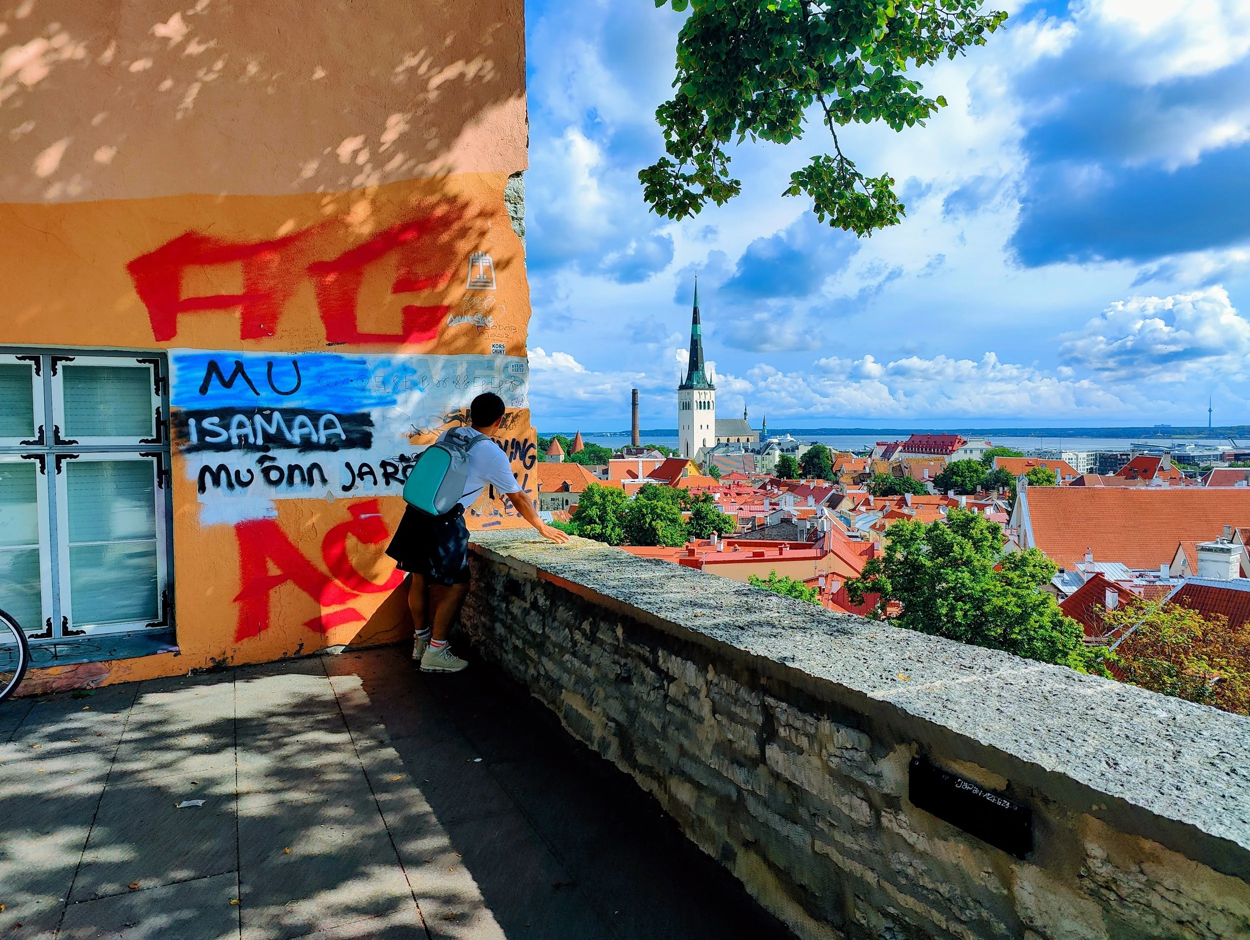 A person leaning on a stone ledge overlooking a cityscape with orange rooftops, a tall church with a green spire, trees, and a distant body of water under a partly cloudy sky. The wall behind the person has colorful graffiti art.