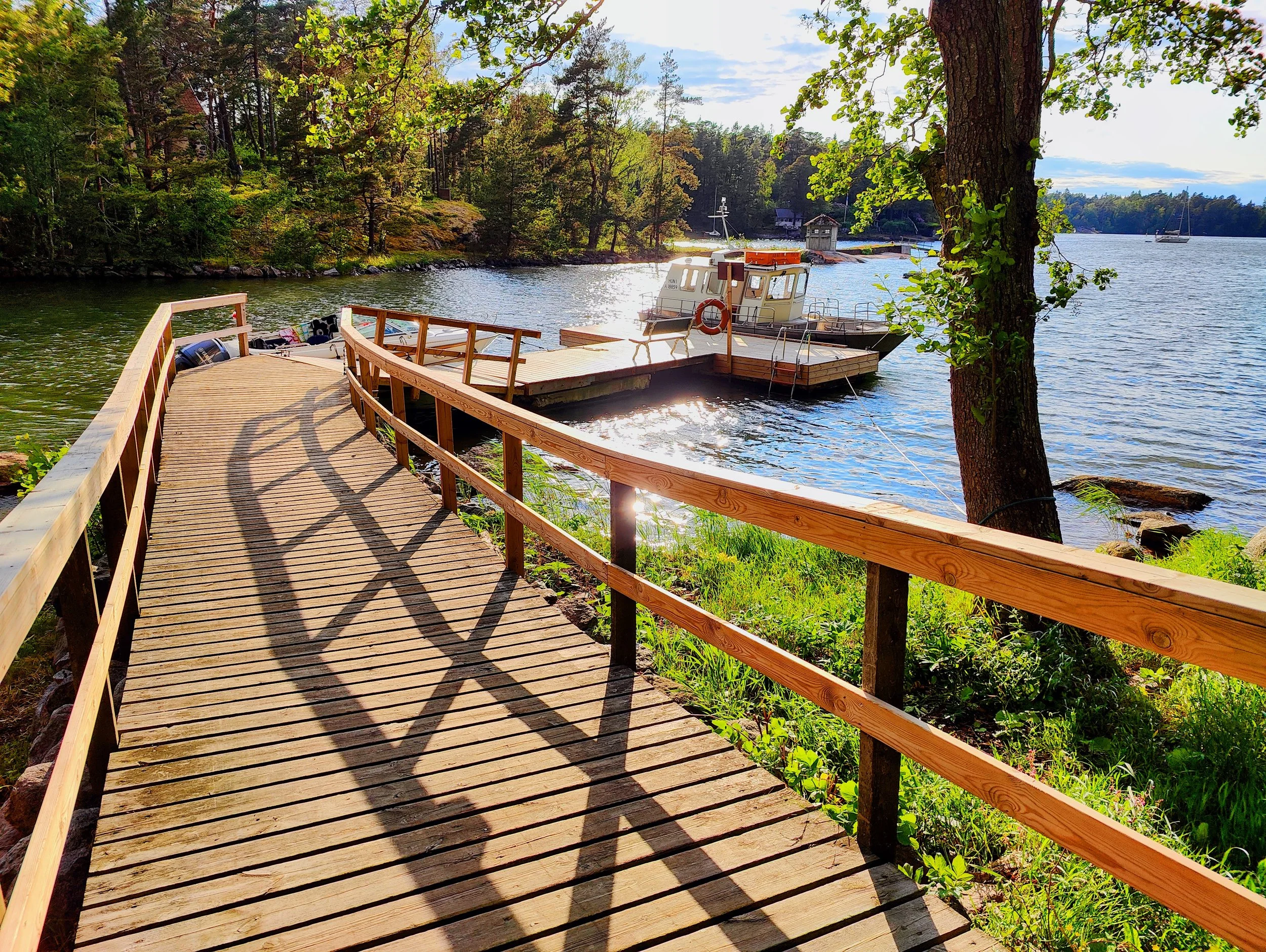 Wooden boardwalk curving along a lakeshore with a dock and boat tied to it, surrounded by trees and greenery, during sunlight.