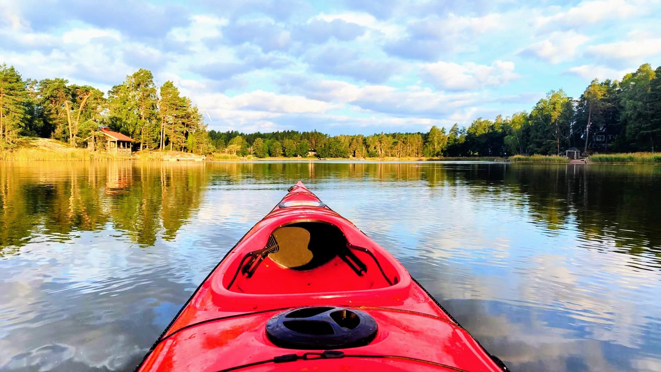 View from a red kayak paddling on a calm lake with lush green trees and houses along the shoreline under a partly cloudy sky.