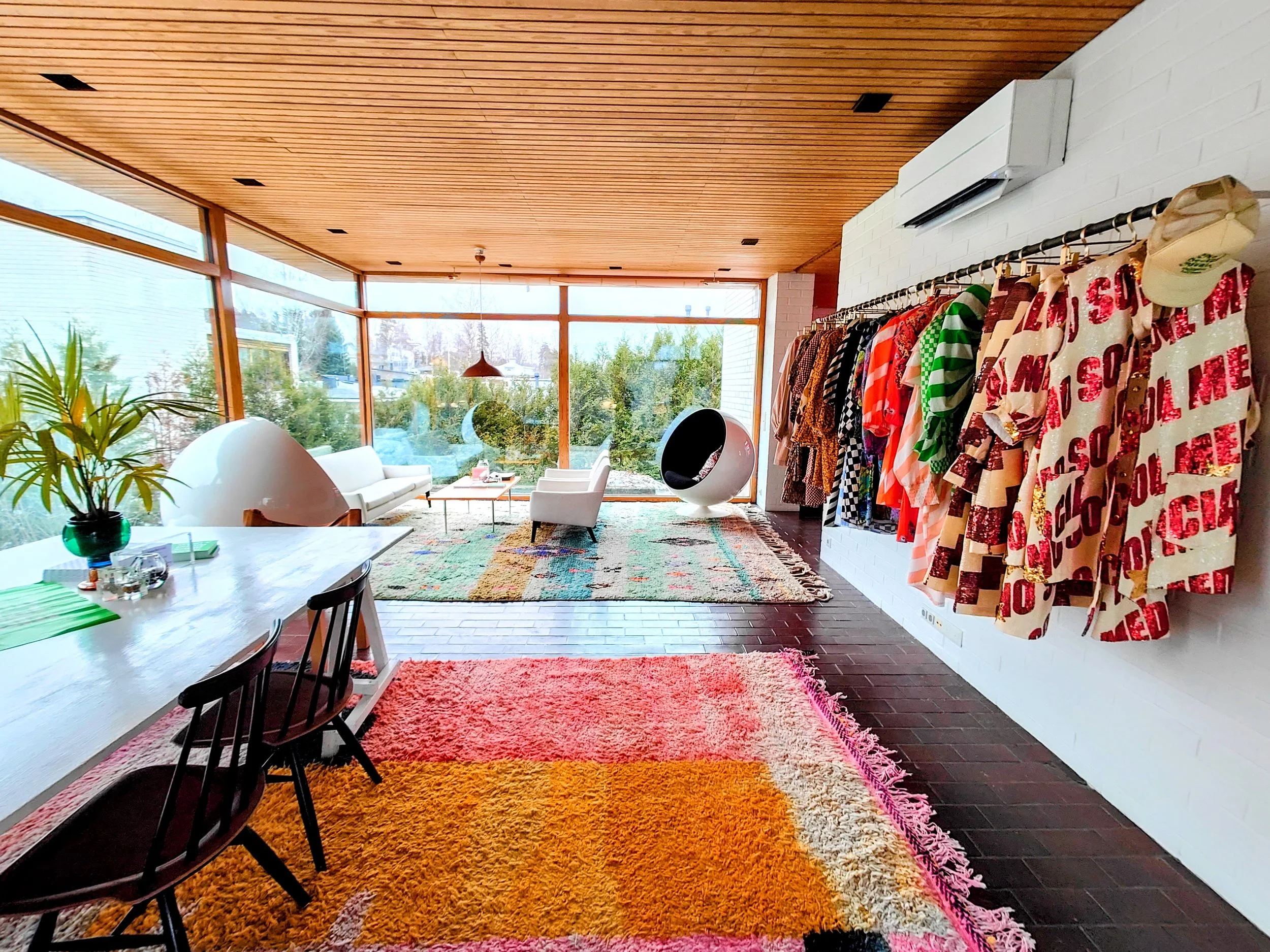 Bright, modern room with large windows, colorful rugs, clothing rack with patterned garments, and contemporary furniture.