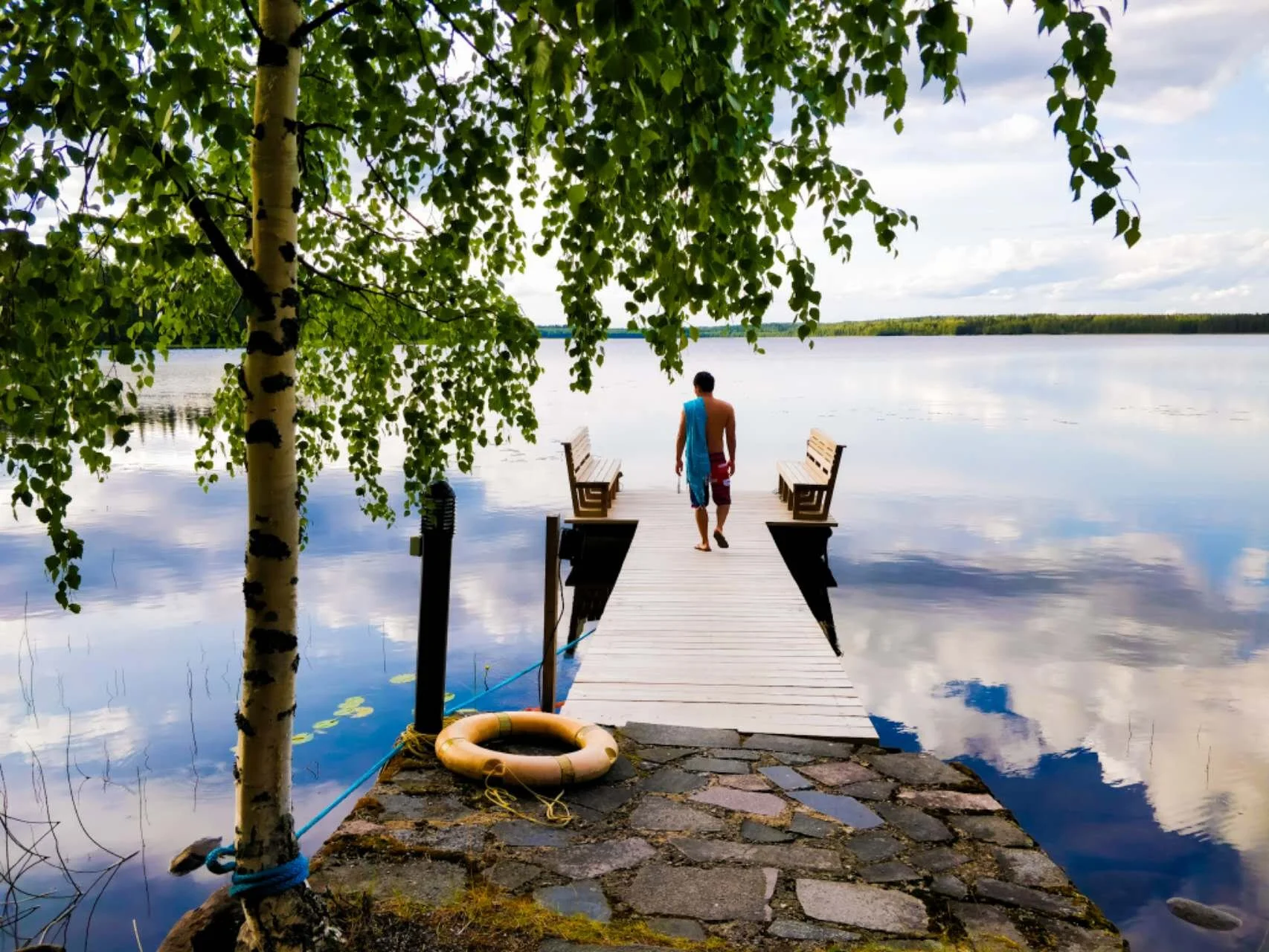 A person in red shorts and a blue towel walking on a wooden pier toward a calm lake, with benches on either side and trees overhead, reflecting the cloudy sky.