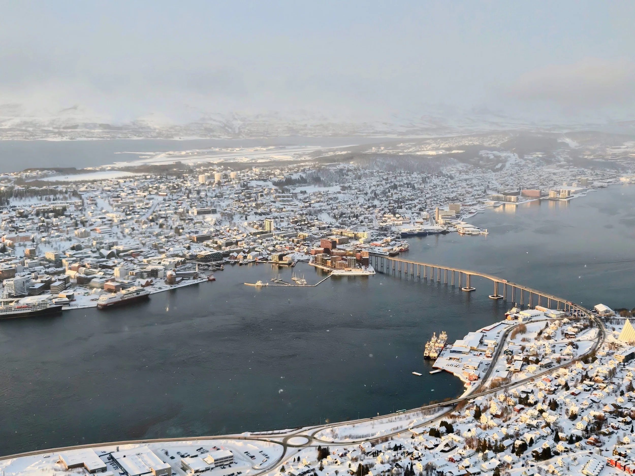 Aerial view of a snow-covered city by a river with a bridge, ships, and numerous buildings during winter.