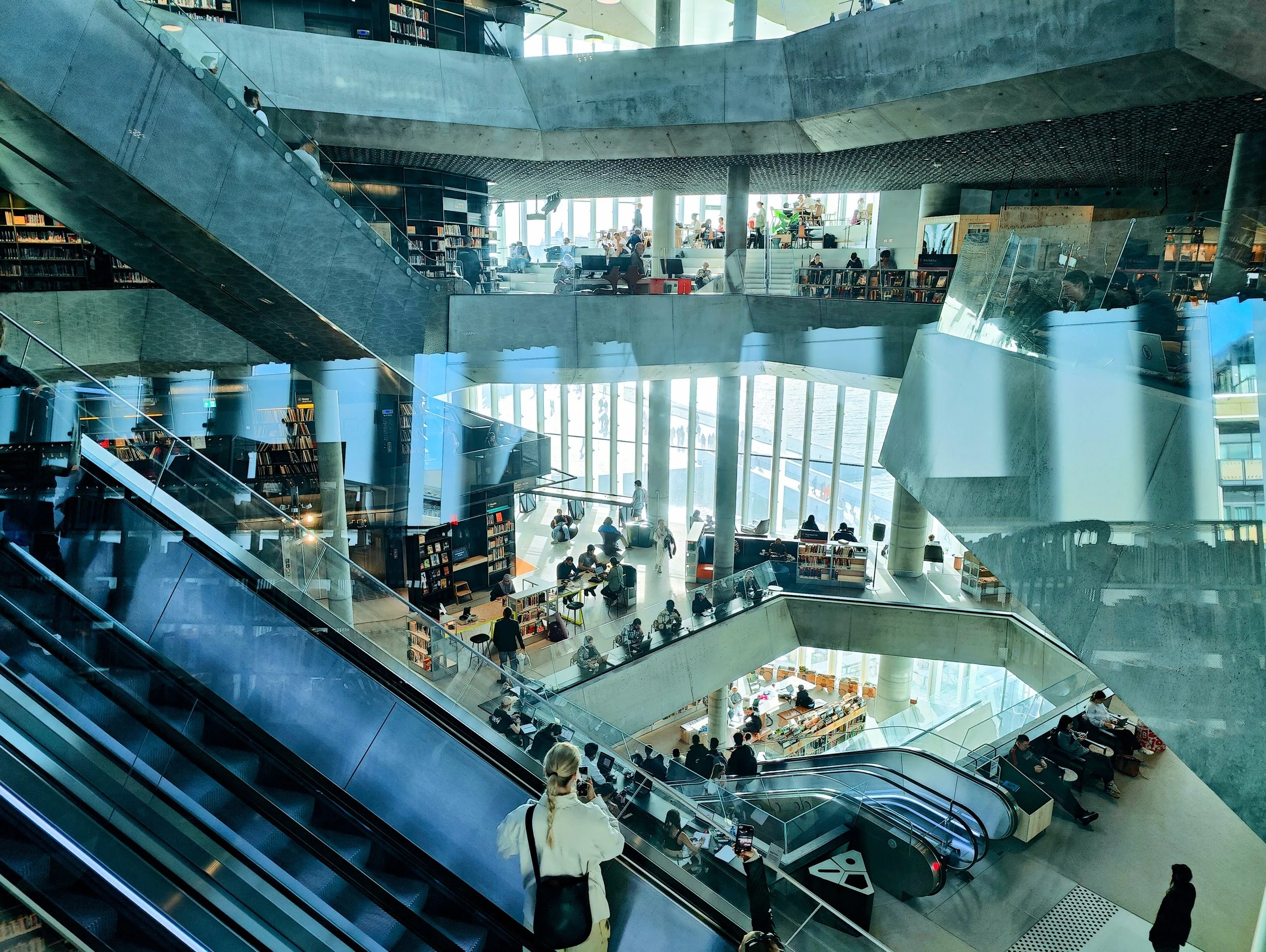 Interior of a modern multi-level library with bookshelves, people reading, working, and walking, featuring escalators, large glass windows, and concrete architecture.