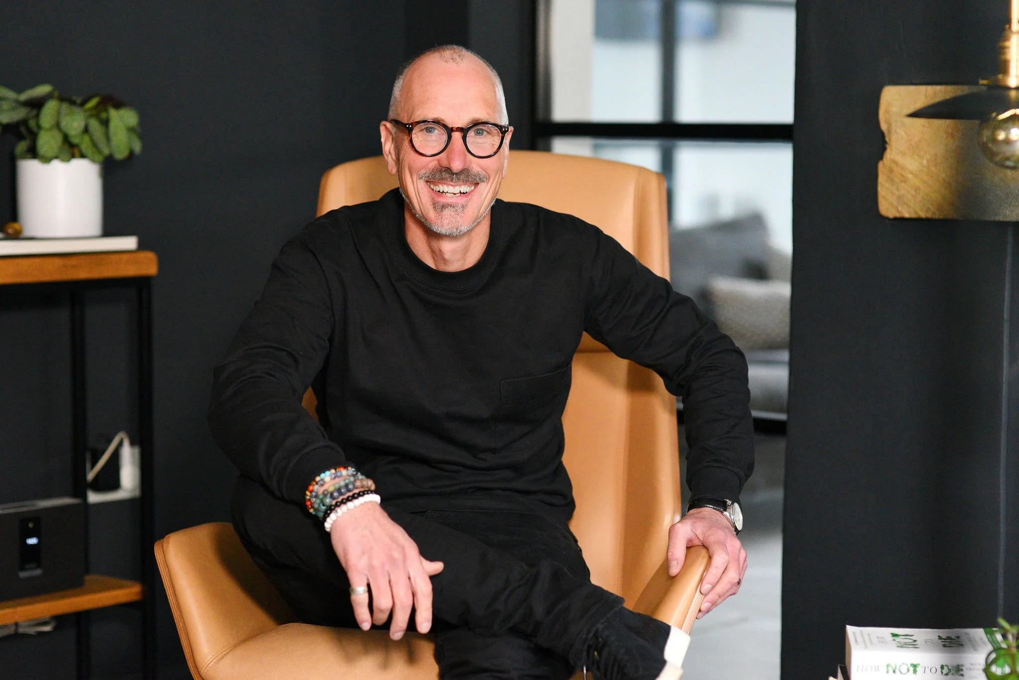 A smiling middle-aged man with glasses, wearing a black long sleeve shirt and black pants, sitting on a tan leather chair in a modern office with dark walls, a window, a desk with a potted plant, and books.