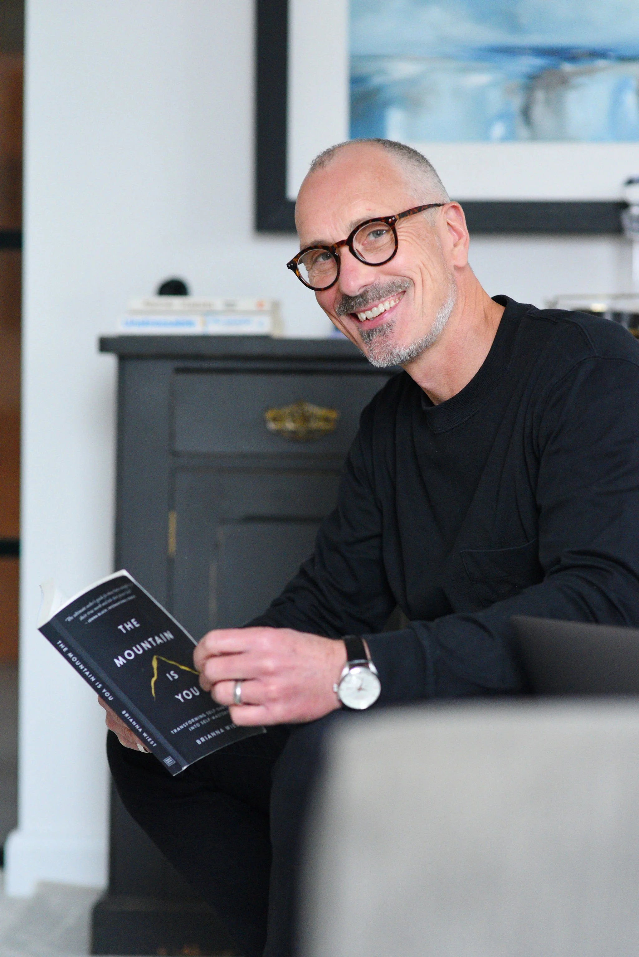 Smiling man with glasses sitting on a couch holding a book titled 'The Mountain is You' in a living room.