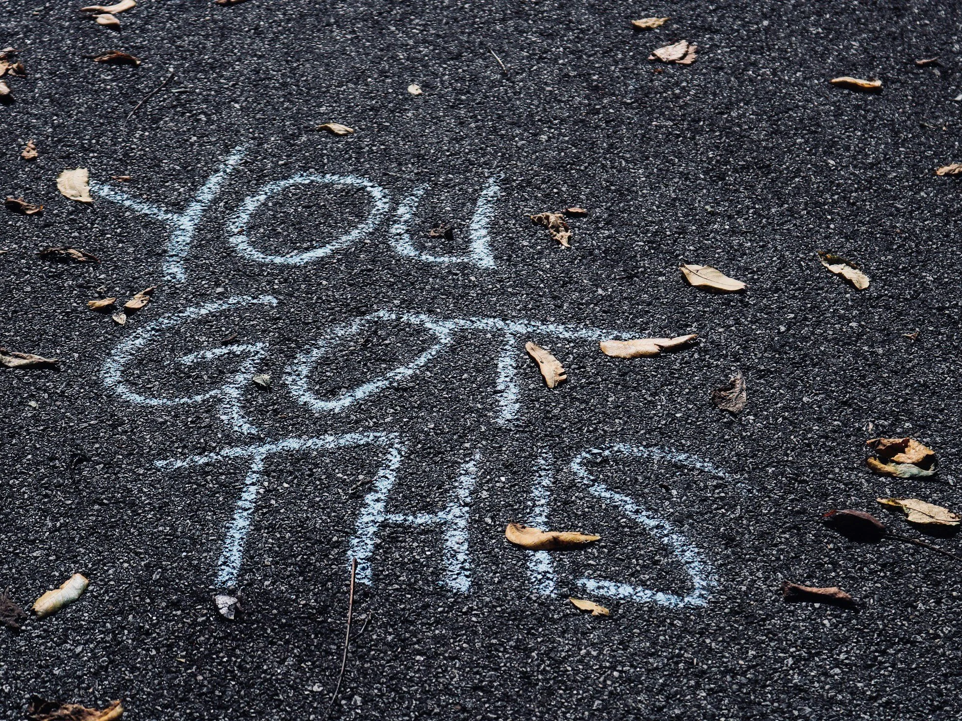 Words written in white chalk on an asphalt ground say "You are loved." scattered dry leaves also on the ground.