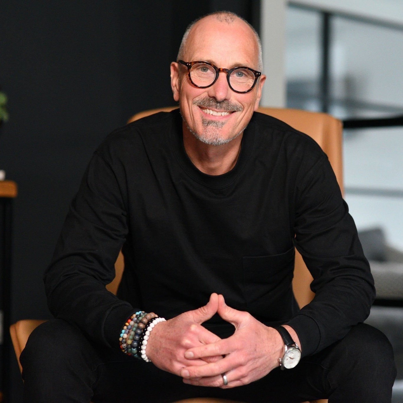 Portrait of a smiling man with glasses, wearing a black shirt, sitting in a chair indoors.