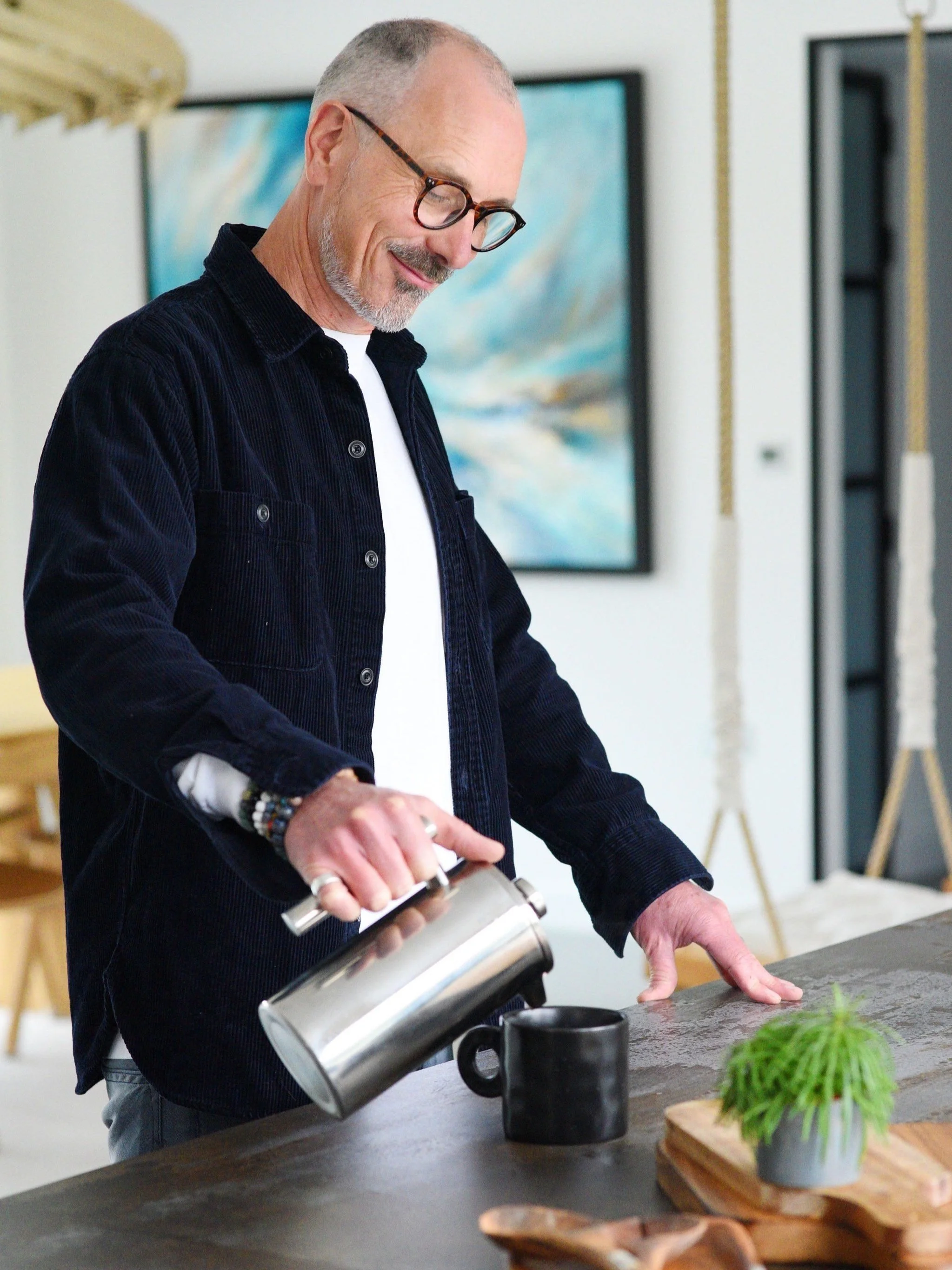 A man with glasses pouring coffee into a black mug on a wooden table with a small green potted plant and wooden tray, in a modern home interior.