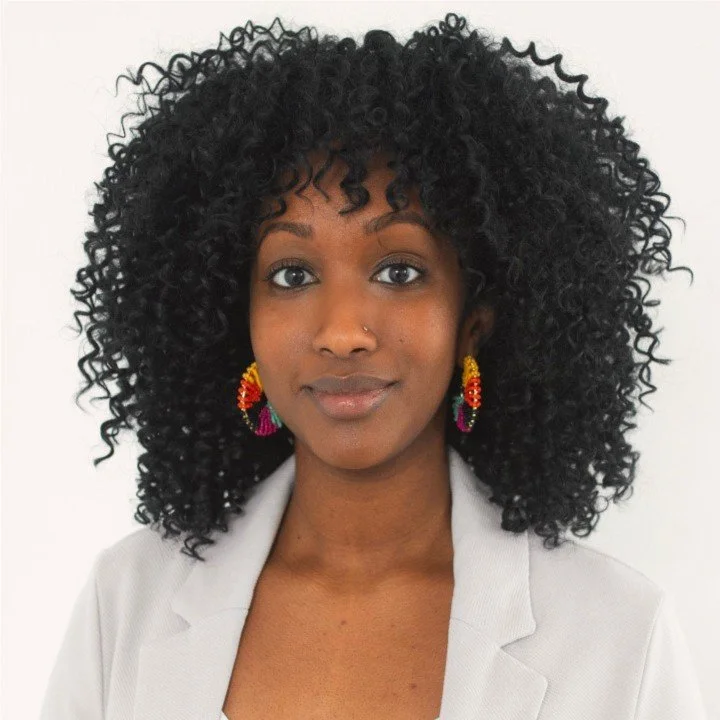 A woman with dark skin and curly black hair wearing colorful earrings and a light-colored blazer against a white background.