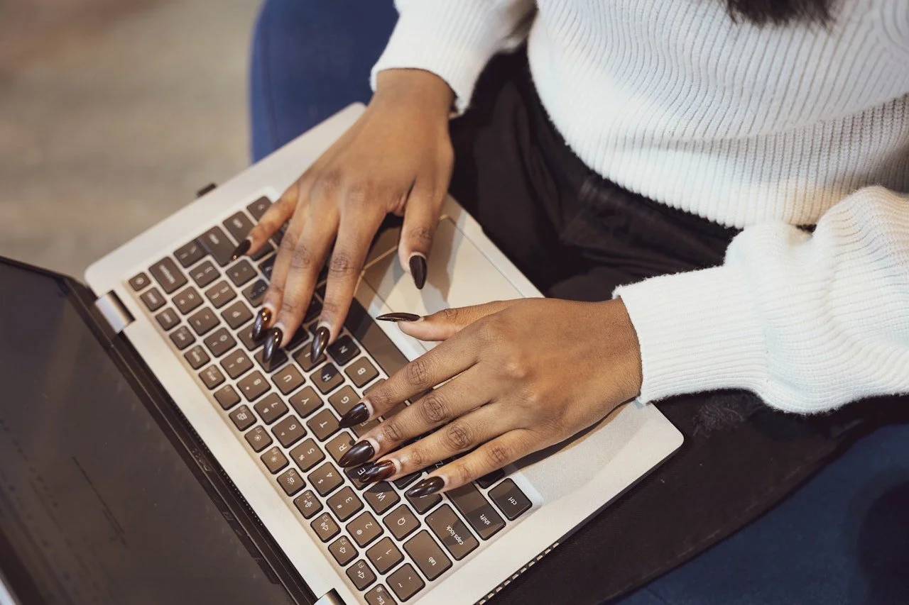 Person typing on a silver laptop keyboard, wearing a white sweater and having black nails.