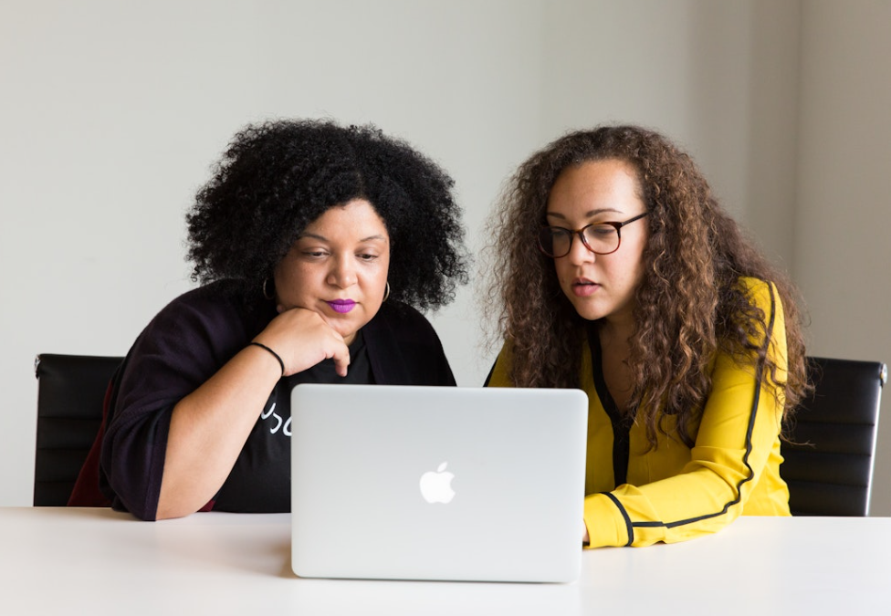 Two women sitting at a table, looking at a laptop, in a plain room.