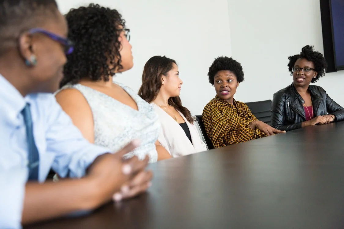 Five women of diverse backgrounds seated at a conference table in a professional setting, engaged in a discussion.