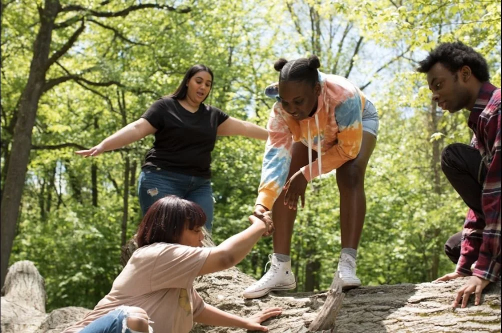 Group of five young adults outdoors in a forest, with one person helping another across a fallen log.