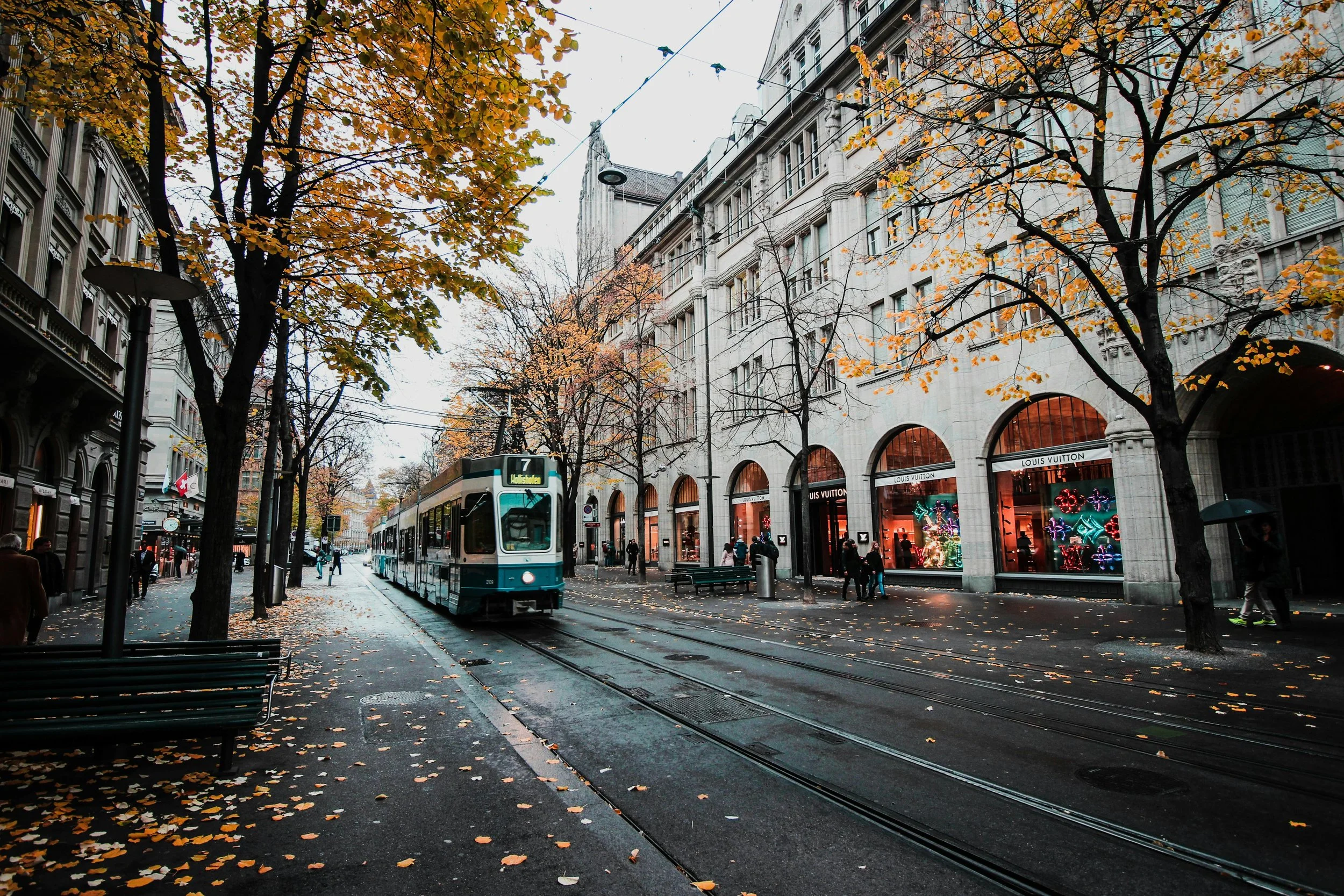 City street scene in autumn with trees losing leaves, a tram on tracks, and shoppers next to a Louis Vuitton store.