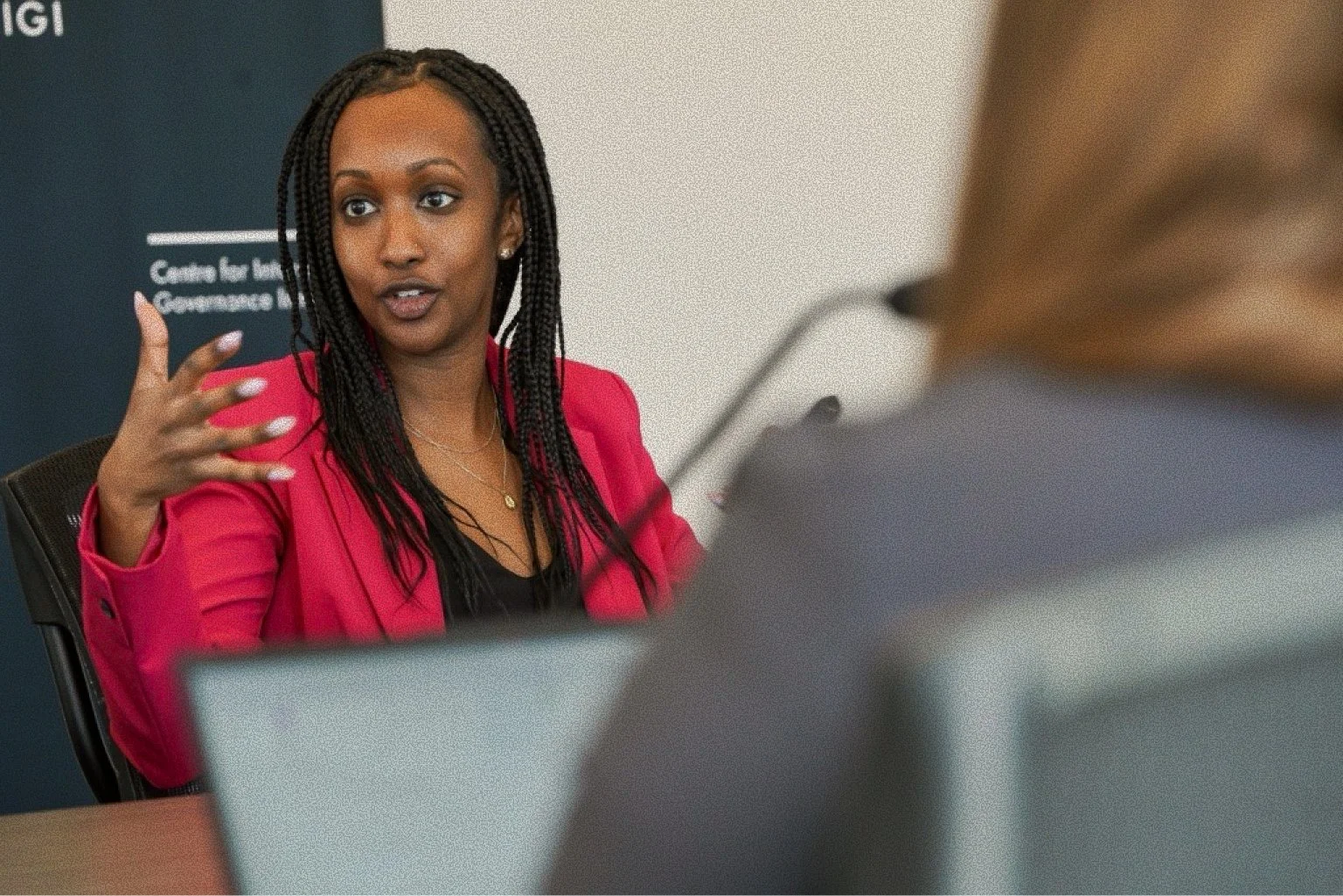 A woman with braided hair in a pink blazer is speaking during a meeting or interview, gesturing with her hand. Part of another person's shoulder and back of their head are visible in the foreground.