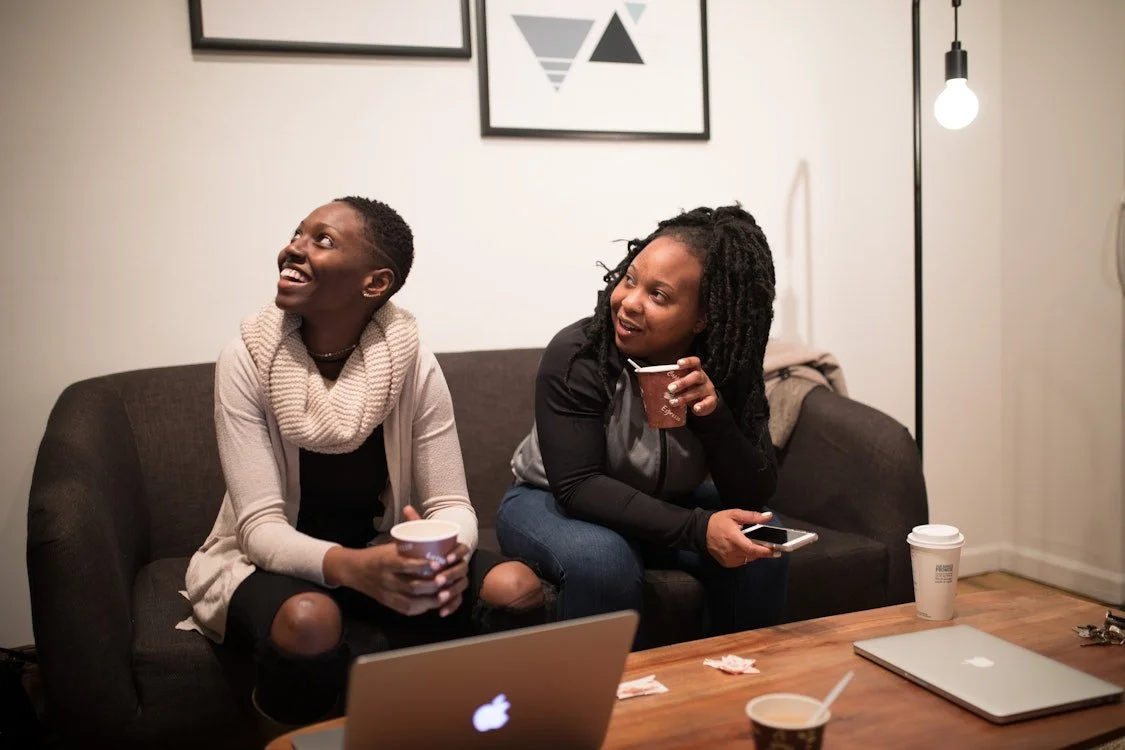 Two women sitting on a dark gray couch, smiling and talking, each holding a coffee cup, in a casual living room with artwork on the wall and a wooden coffee table with laptops, cups, and a cellphone.