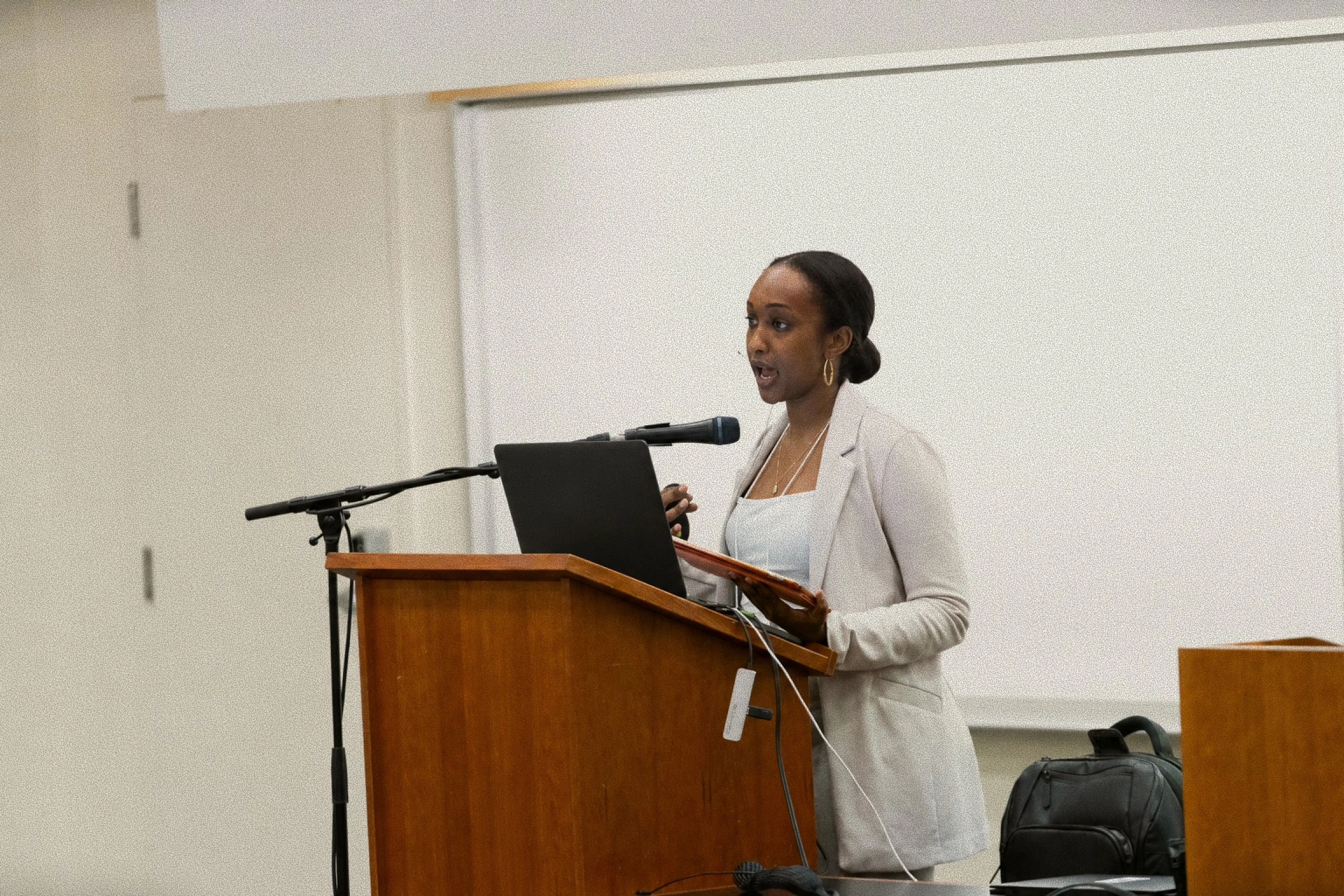 African American woman with dark hair tied back, wearing earrings and a light-colored blazer, speaking at a podium with a laptop, in a room with a whiteboard.