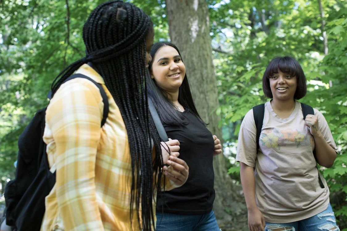 Three young women with backpacks talking and smiling in a wooded area.
