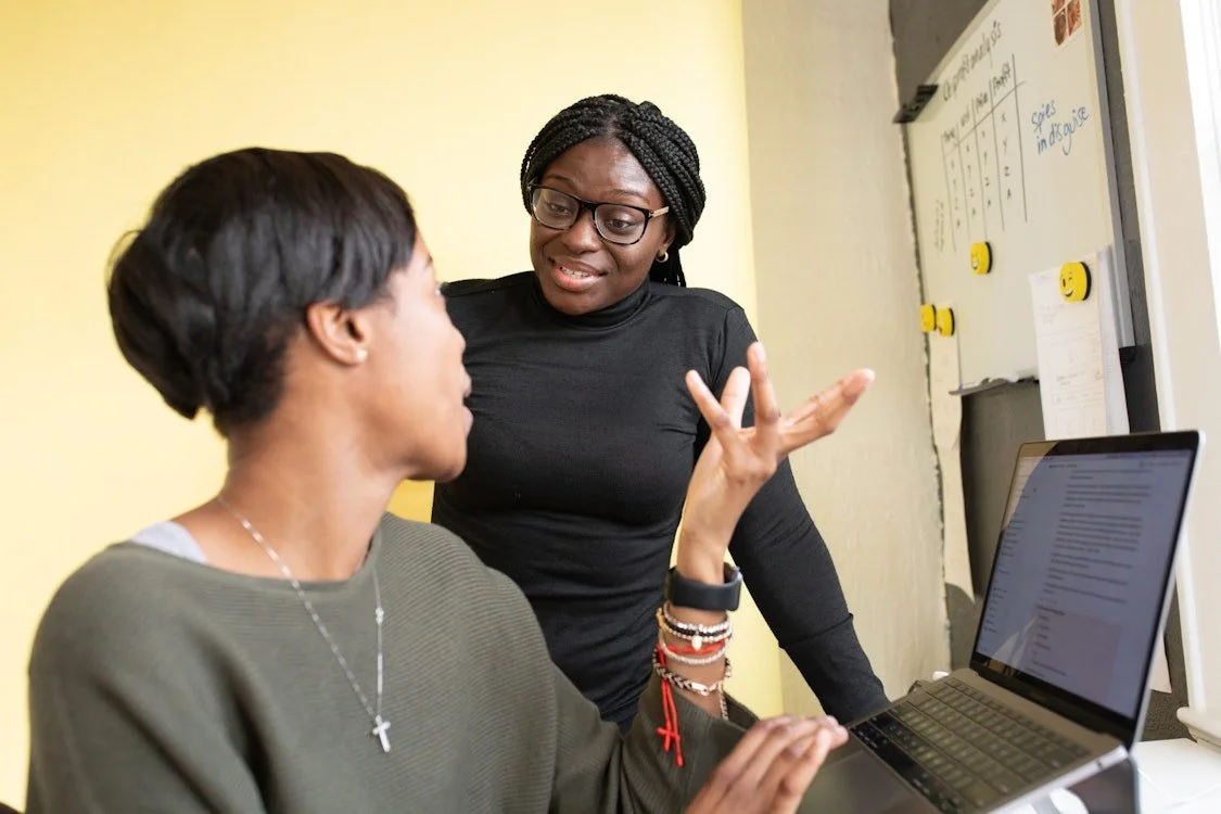 Two women having a serious conversation at a desk, one sitting with a laptop and the other standing with hand gestures.