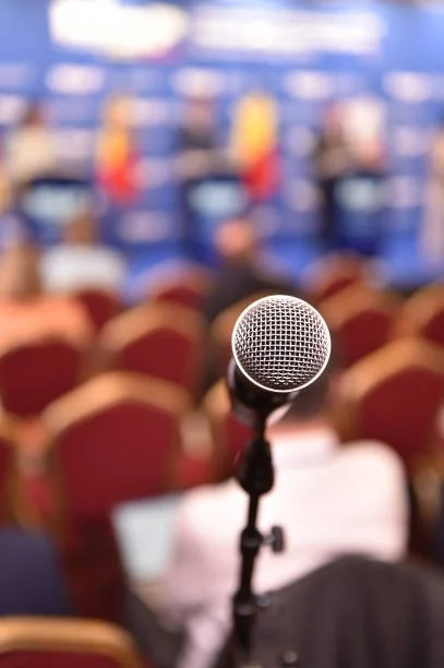 Close-up of a microphone on a stand in a conference room, with blurred chairs and a blue background displaying a large screen.