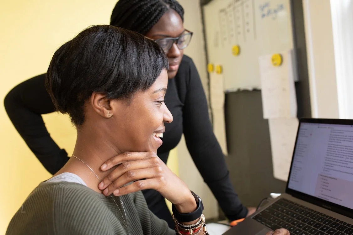 Two women working together at a desk with a laptop, one woman smiling and the other leaning over to look at the screen.