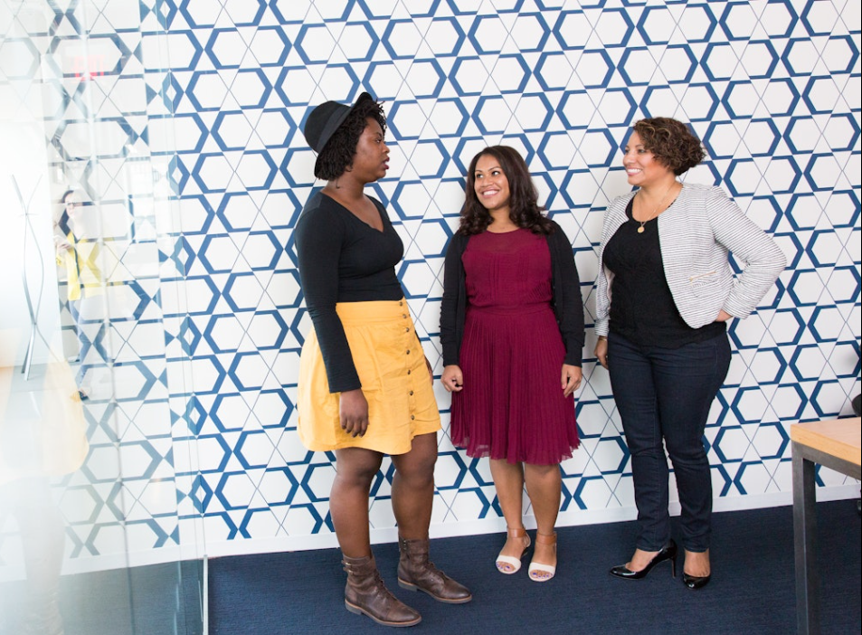 Three women having a conversation in an office with a blue geometric patterned wall.