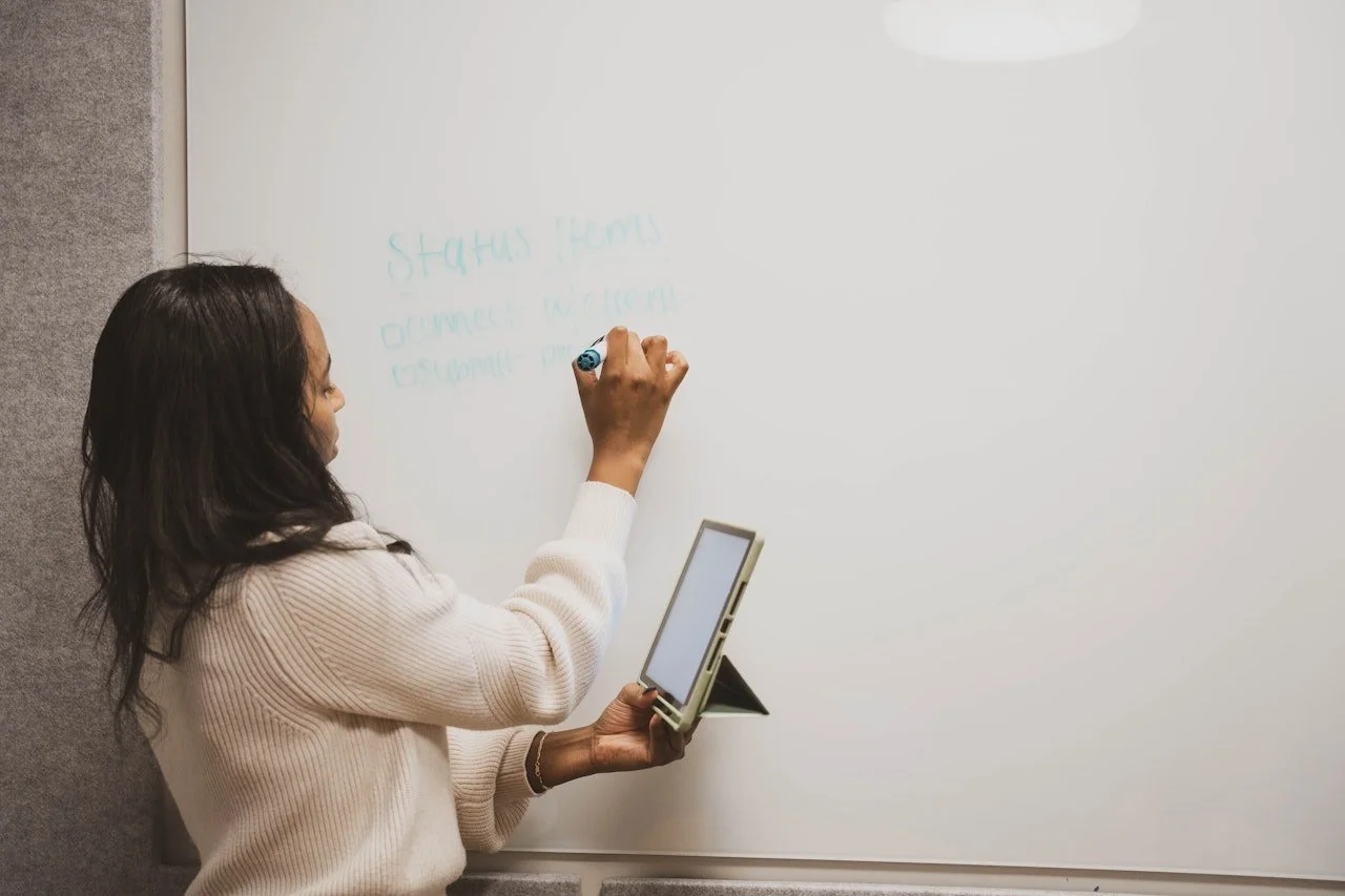 Woman writing on a whiteboard while holding a tablet in her left hand.