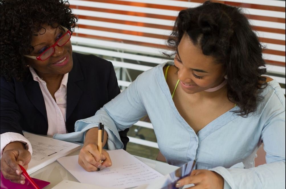 An older woman with curly hair, glasses, and a black blazer is smiling while sitting next to a young woman with curly hair, a white shirt, and a green strap, both are looking at some documents and writing.