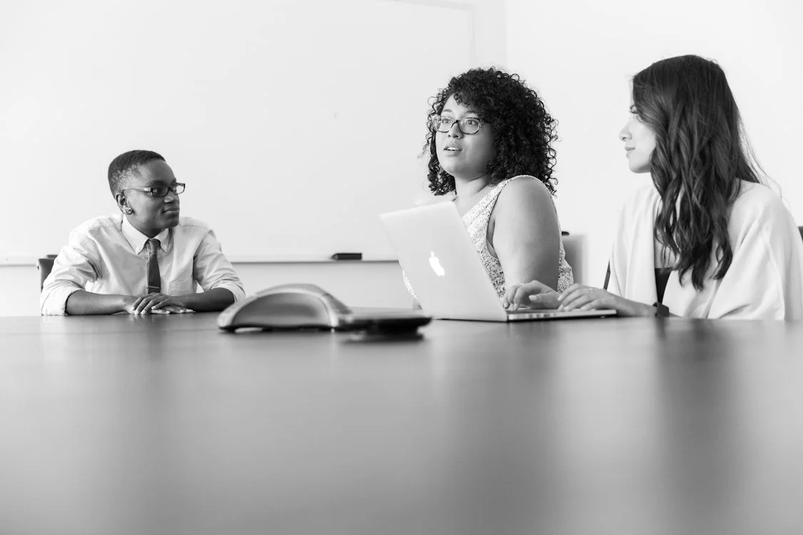 Three women sitting at a conference table in a meeting room, engaged in conversation.