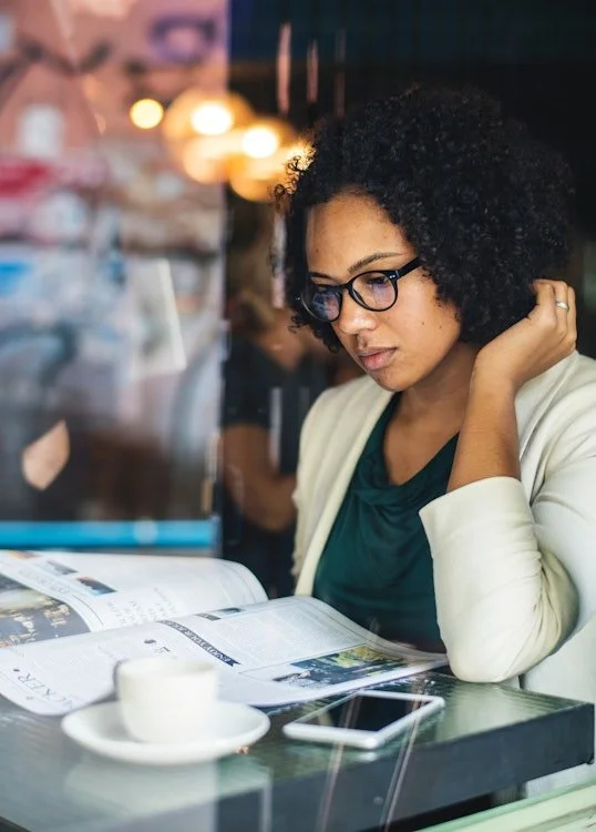 A woman with curly hair and glasses reading a newspaper at a cafe table, with a coffee cup and smartphone nearby, in a lively indoor setting.