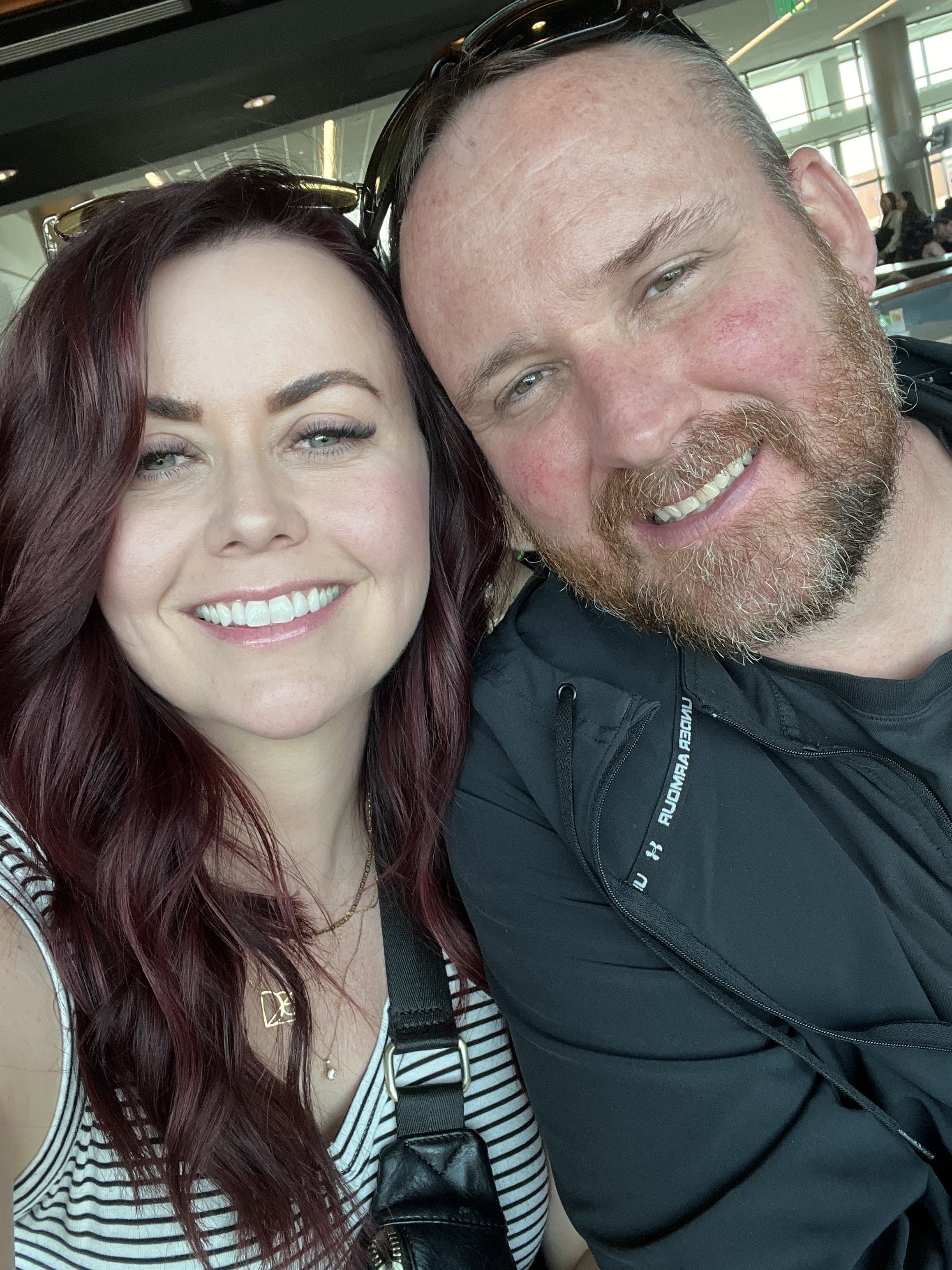 A smiling woman with long red hair and a man with a beard and short hair taking a close-up selfie together indoors.