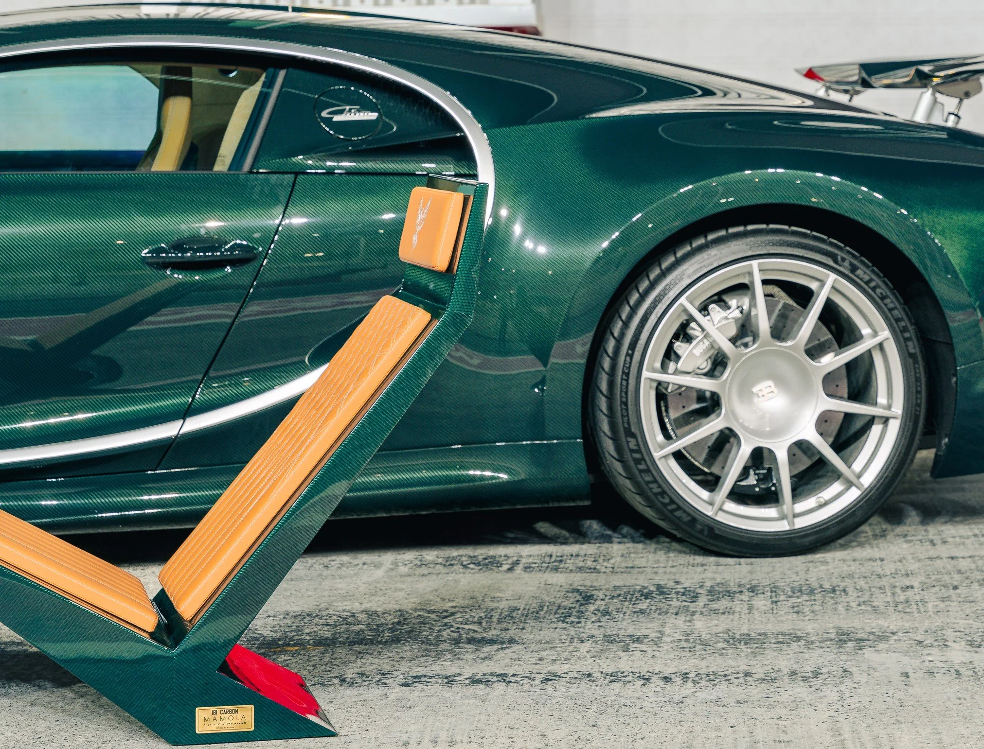 Close-up of a green Bugatti sports car with a carbon fiber finish, showing the rear wheel and part of the side. A tan and green racing seat is positioned next to the car.