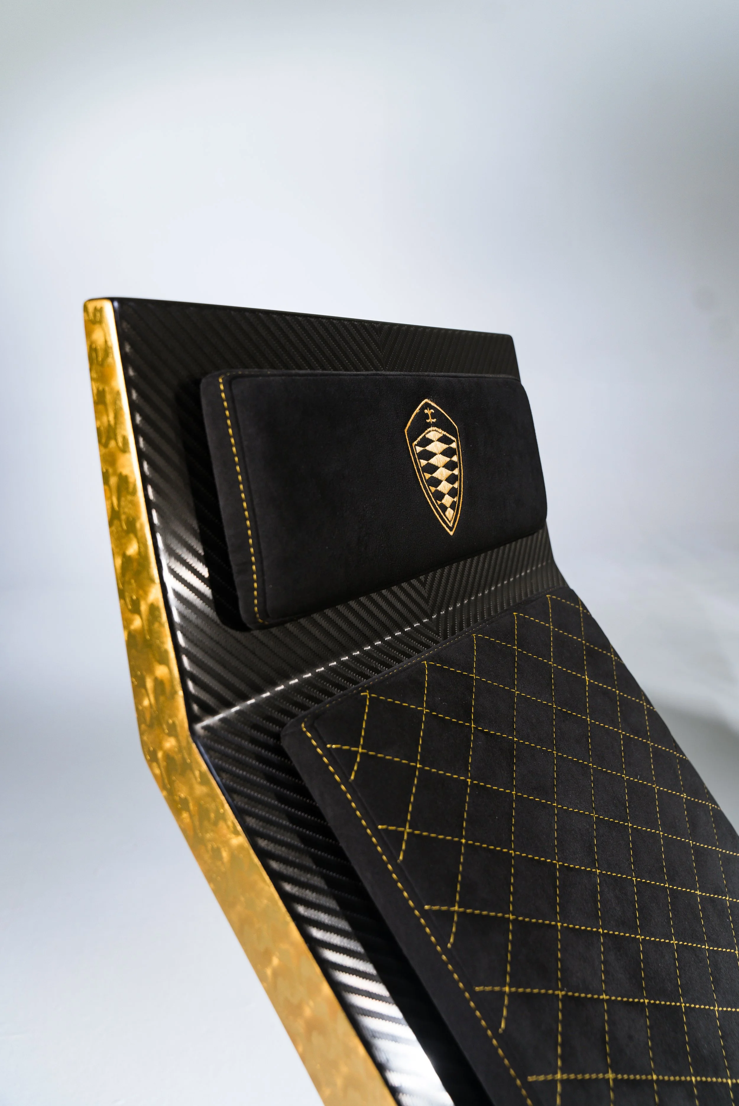 Close-up of a modern, black chair with gold accents, featuring a black cushion embroidered with a checked shield design and a matching quilted black seat.